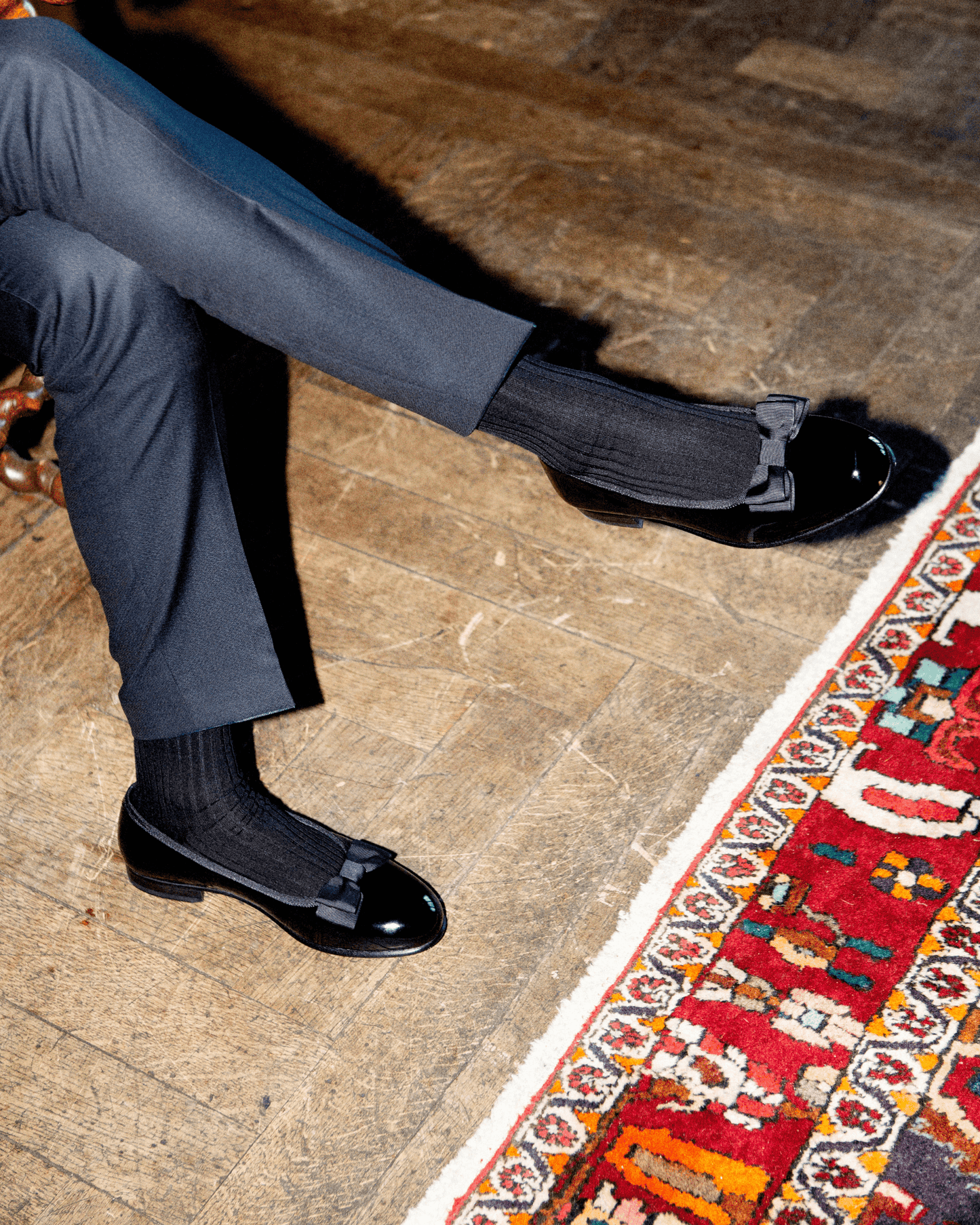 A person seated with legs crossed, wearing Rosendal – Black Patent Leather opera pumps with dark trousers and black socks.