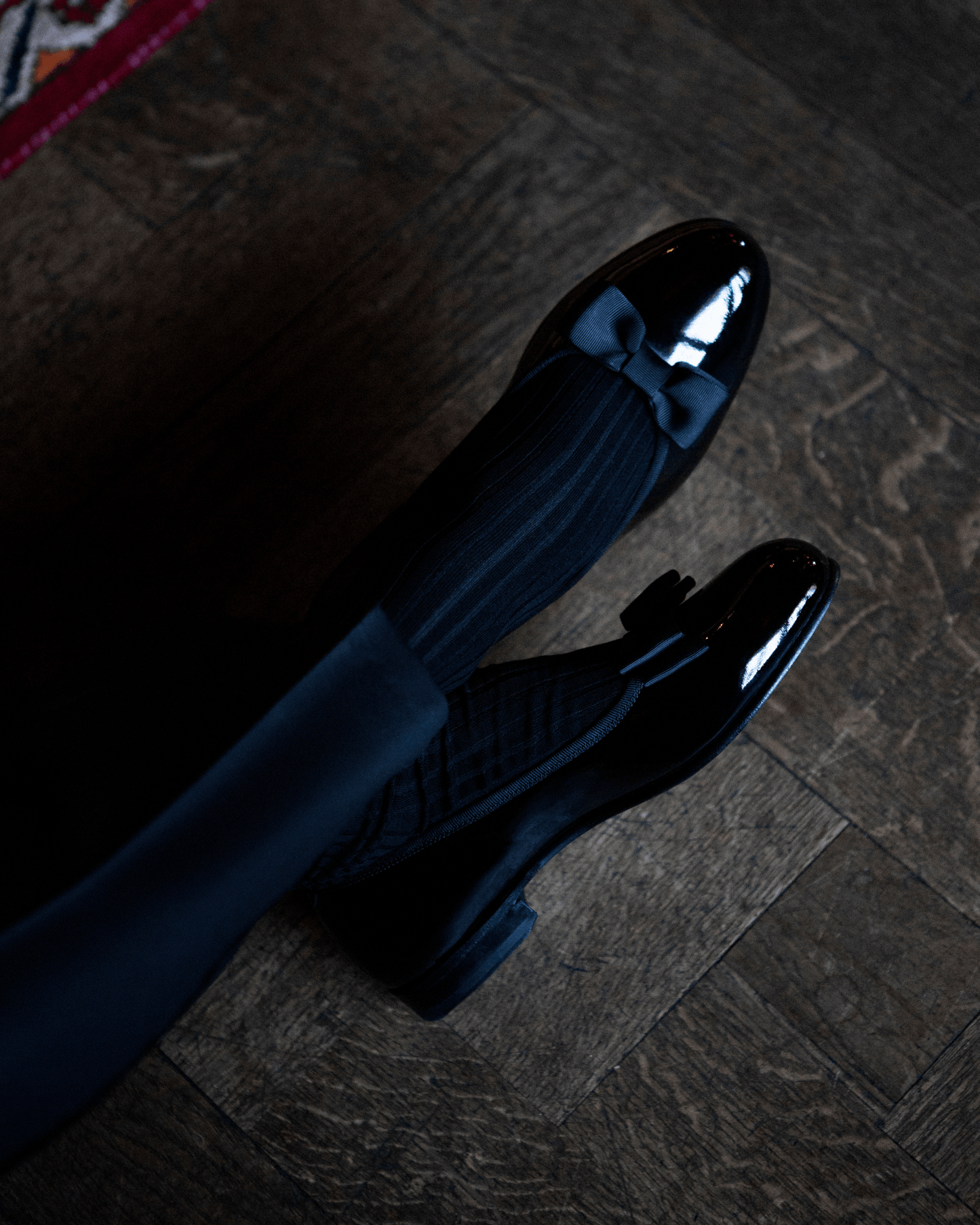 An overhead view of a person wearing Rosendal – Black Patent Leather opera pumps with ribbed socks on a dark wooden floor.