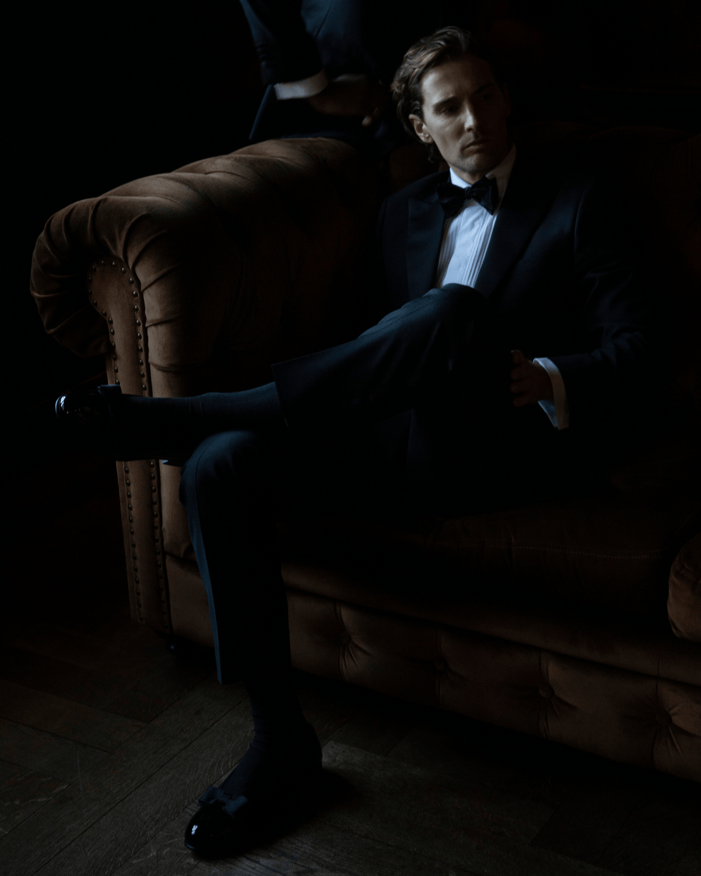 A man in a tuxedo relaxes on a brown tufted sofa, wearing Rosendal – Black Patent Leather opera pumps.