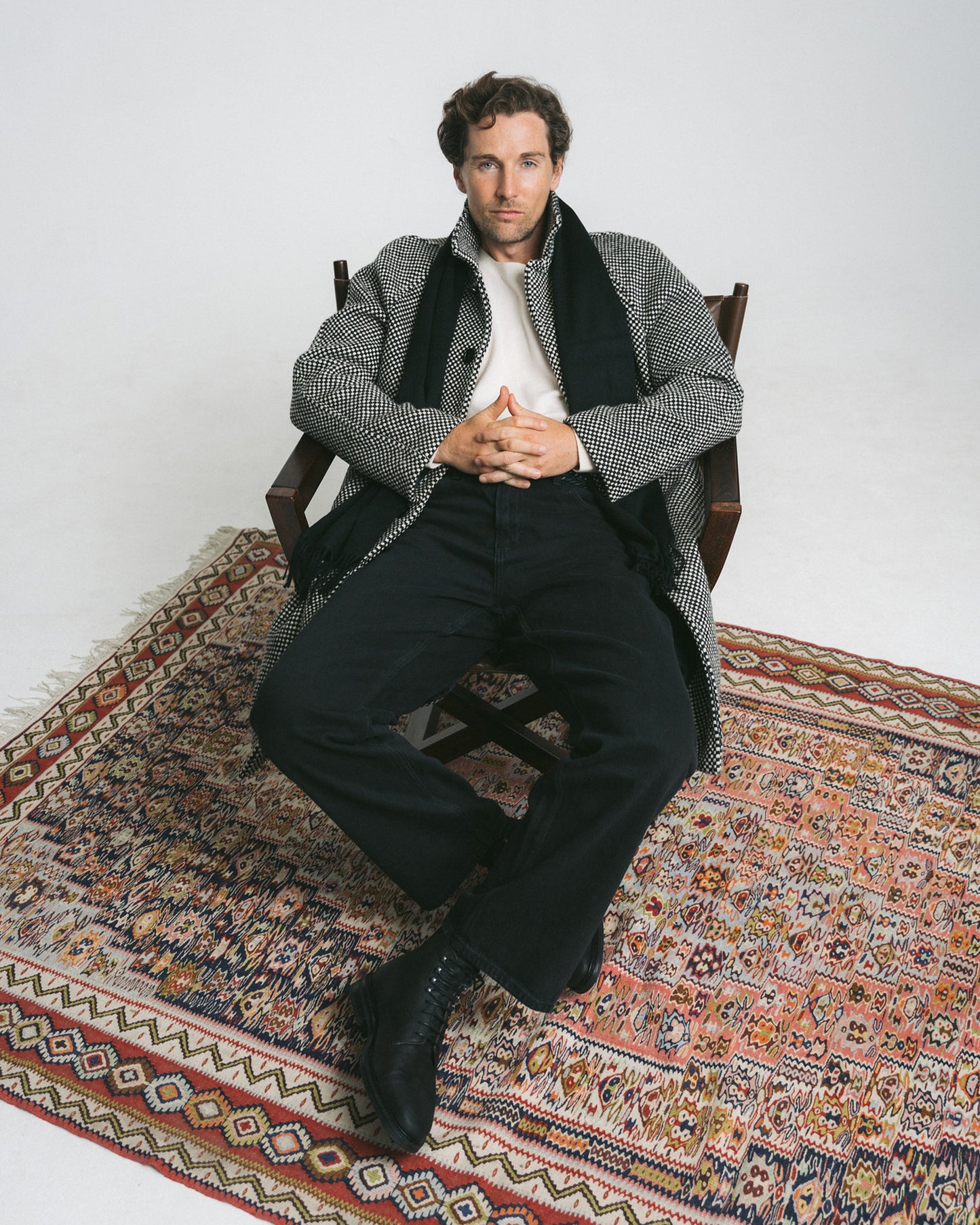 A man sits in a chair on a patterned rug, wearing Abisko – Black Waxed Suede boots with black trousers and a patterned coat.