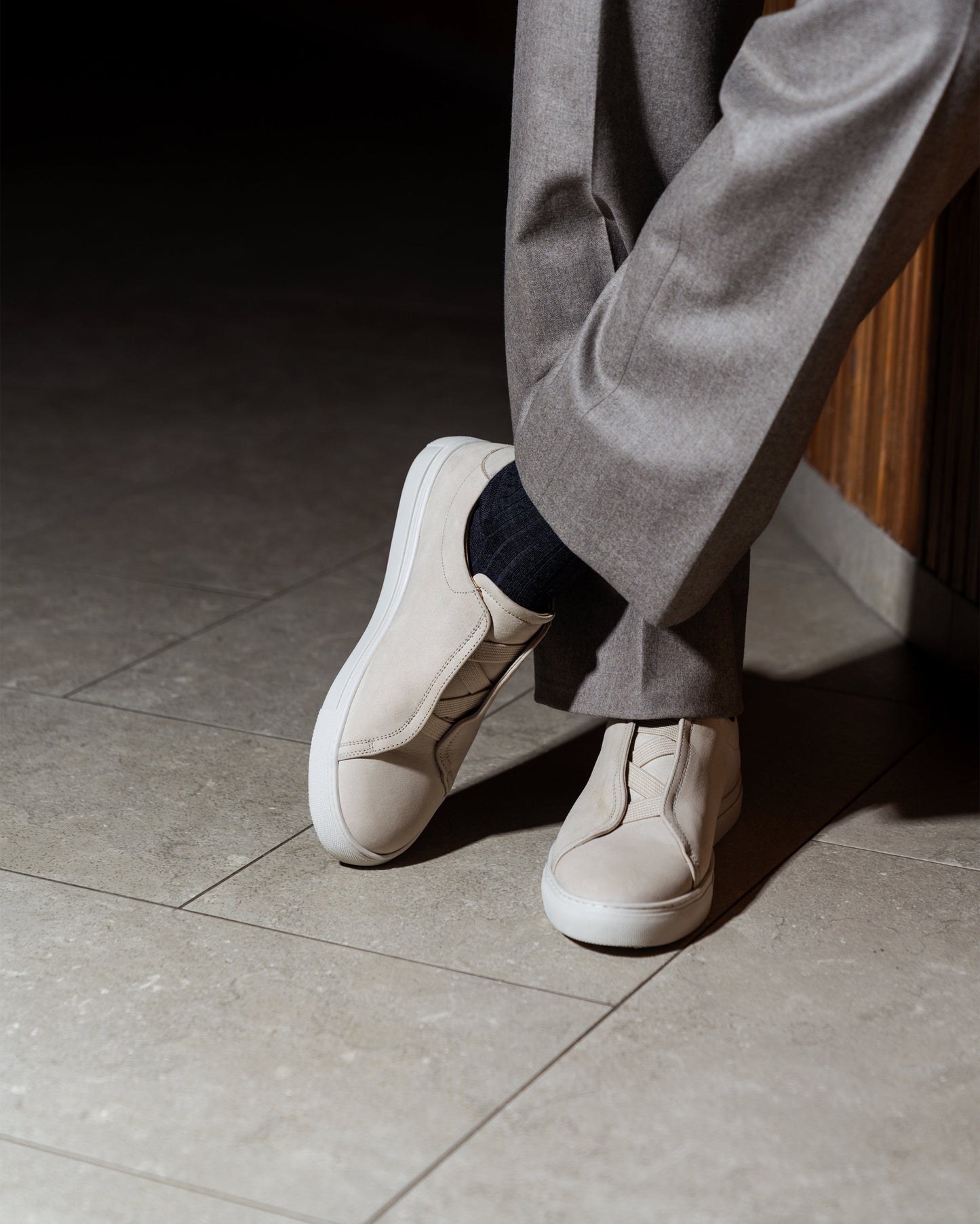 A person wearing Alnö – Beige Nubuck sneakers, styled with light grey tailored trousers and dark socks.