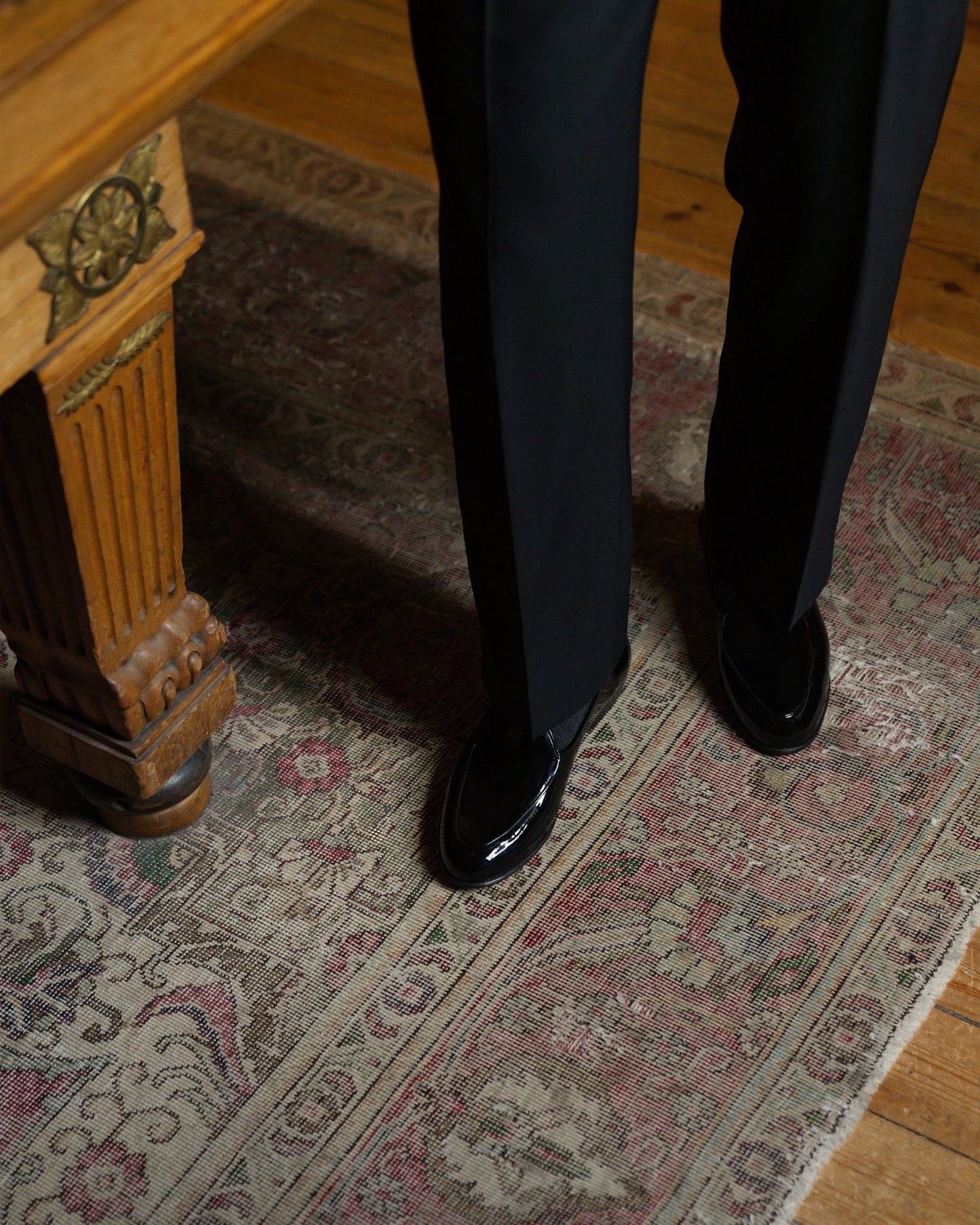 A person wearing Ängby – Black Patent Leather loafers with tuxedo trousers, standing on a vintage-style rug.