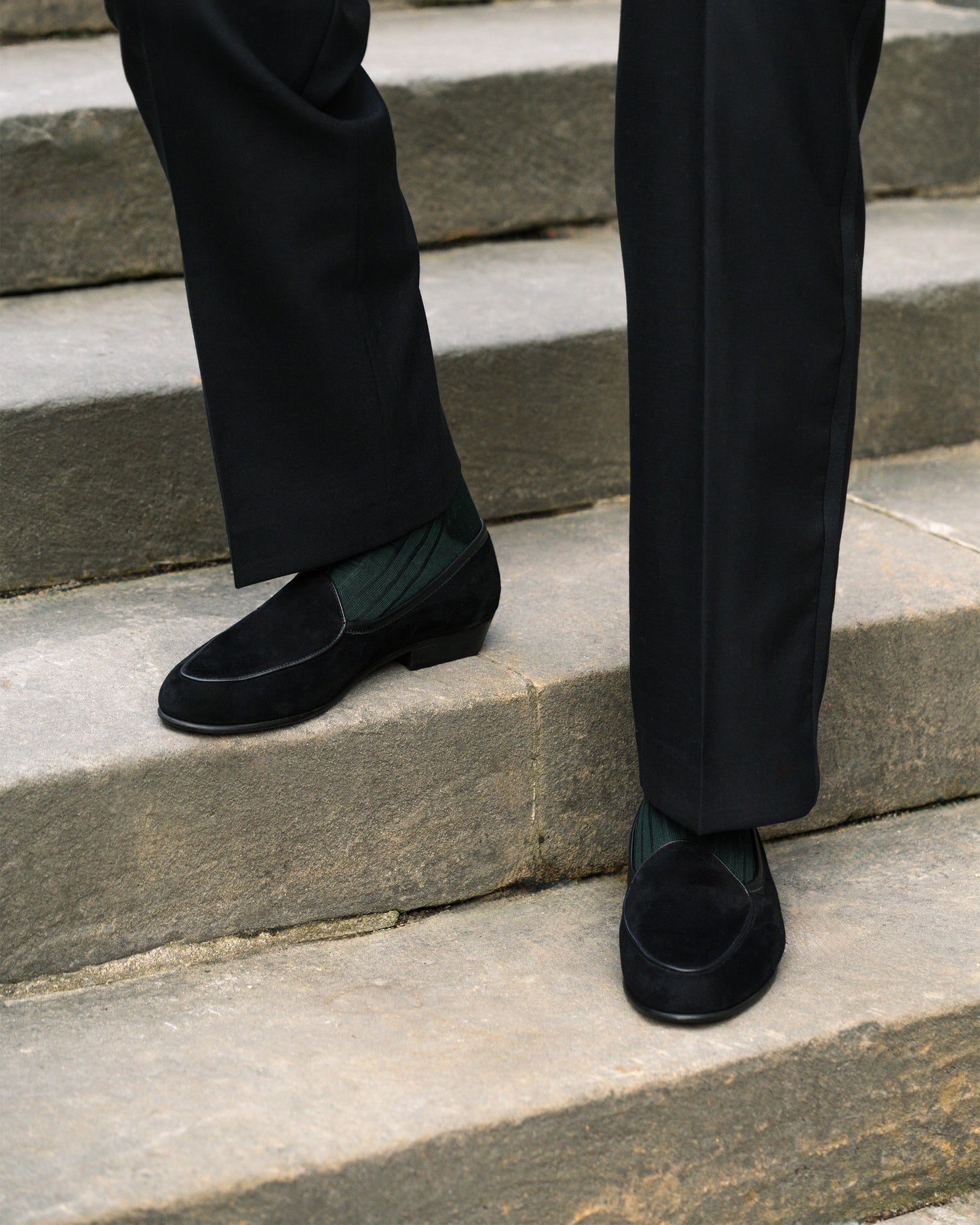 Close-up on a person wearing Ängby – Black Suede loafers with black trousers and dark green socks on stone steps.