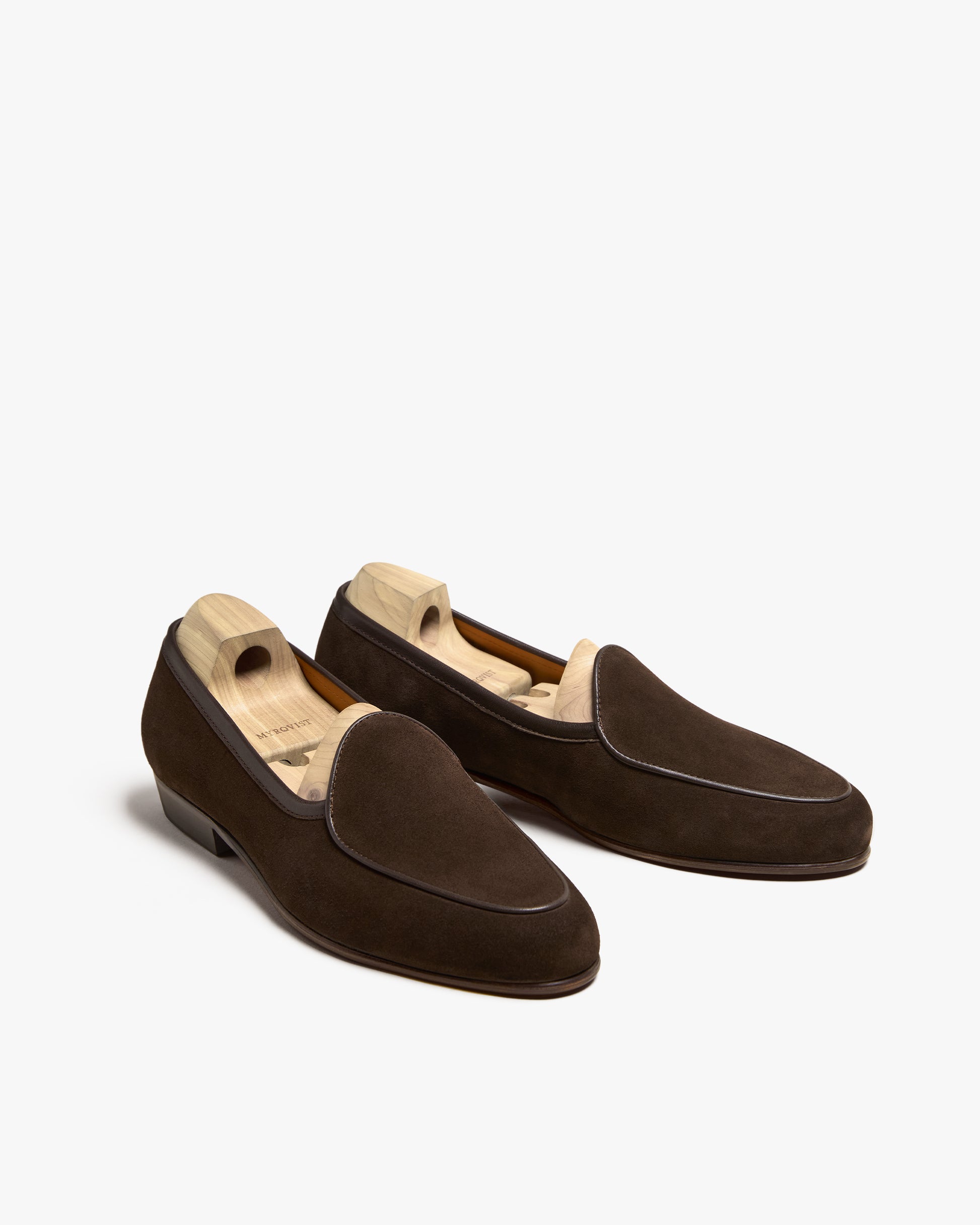 A pair of Ängby – Dark Brown Suede Belgian loafers fitted with wooden shoe trees, on a white background.