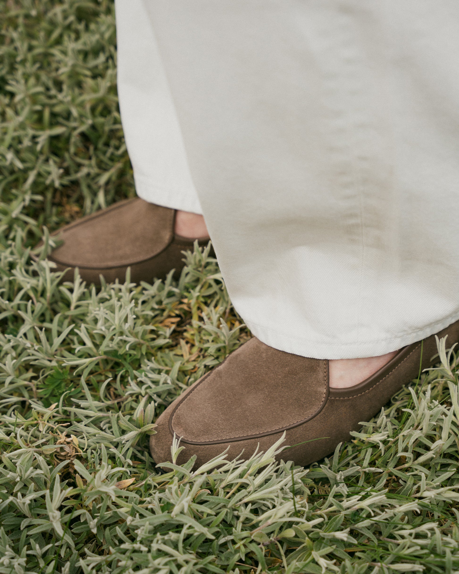 Close-up on a person wearing Ängby – Taupe Suede loafers with white trousers, standing in greenery.