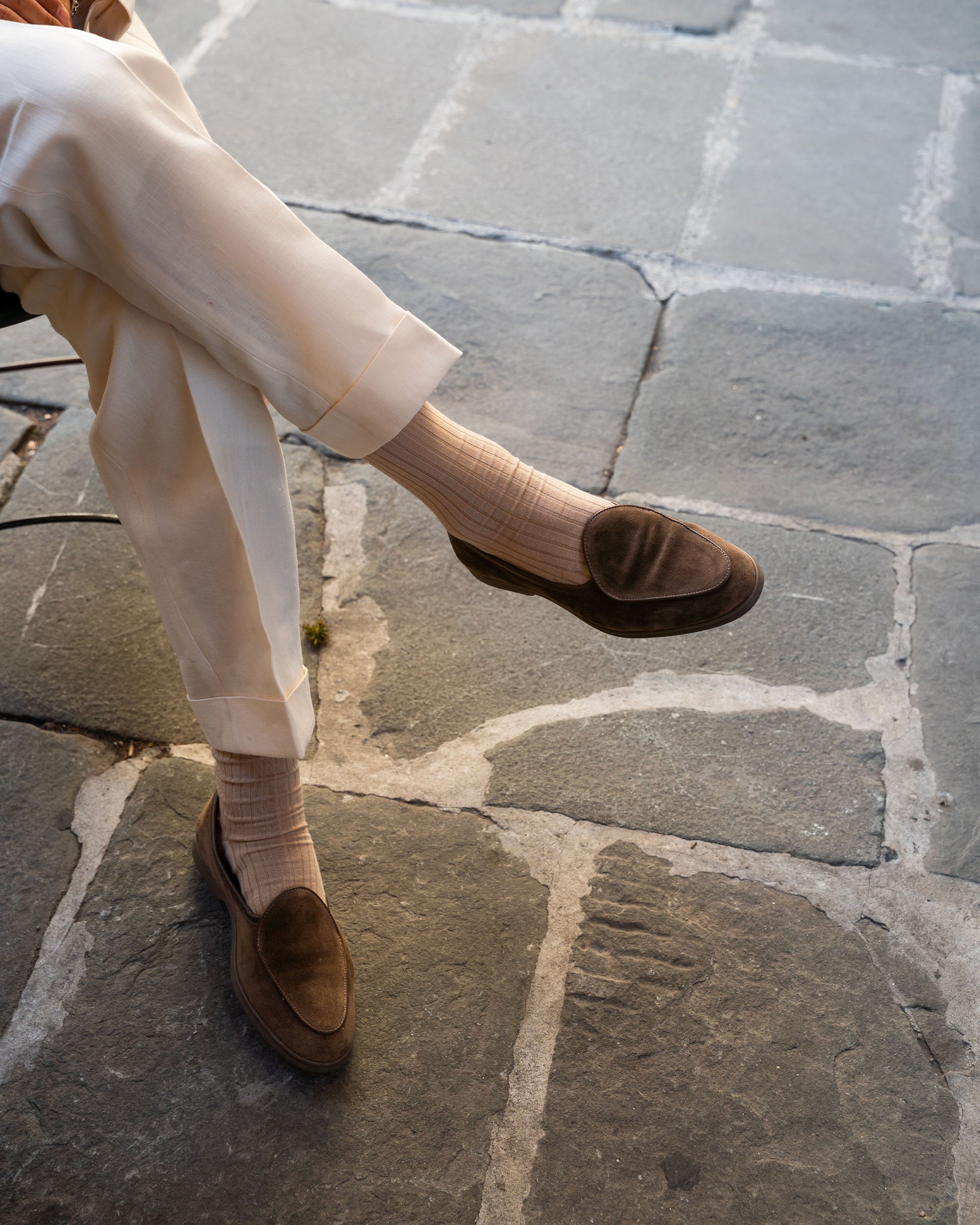 A person sitting with legs crossed on a stone patio, wearing Ängby Walker – Taupe Suede loafers with cream trousers.
