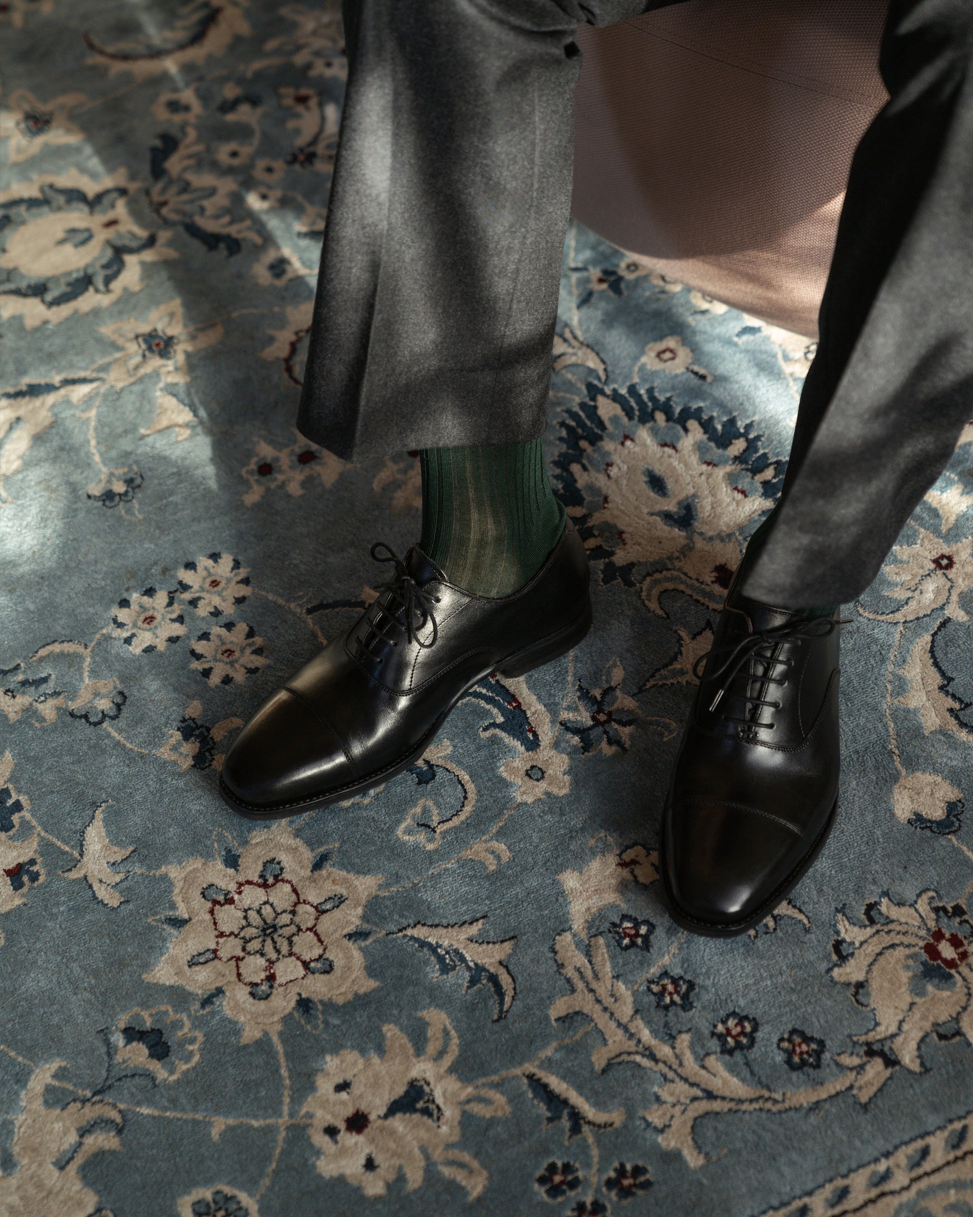 A person wearing Äppelviken – Black Calf Oxfords with grey trousers and dark green socks on a blue patterned rug.