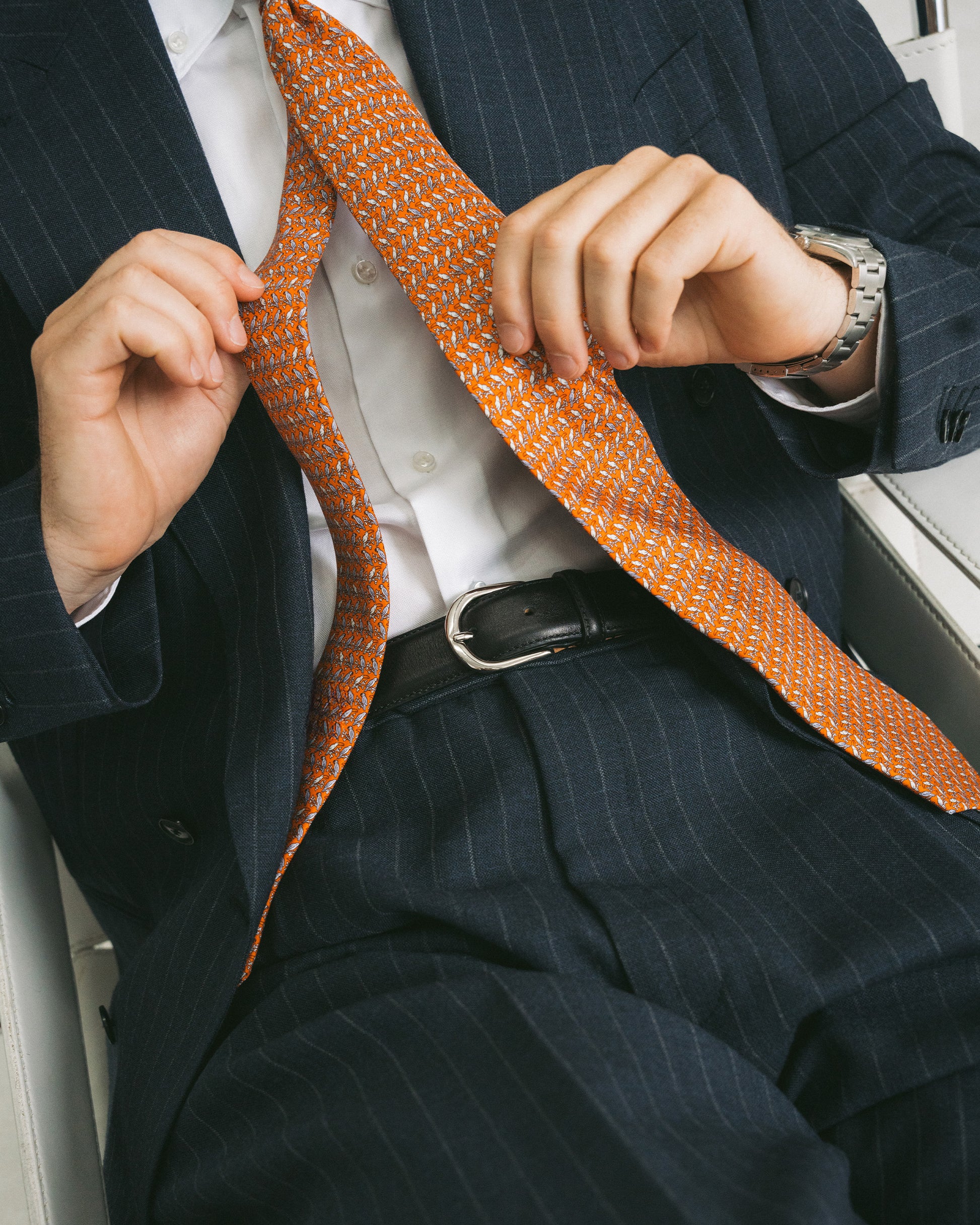 A man in a pinstripe suit adjusting his tie, with the focus on a black leather belt with a silver buckle.