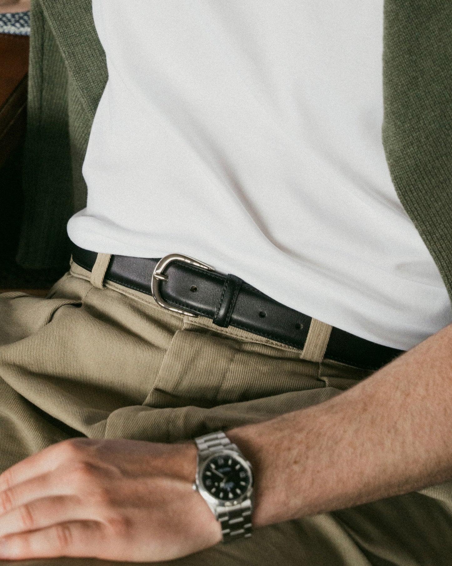 A man styles a black leather belt with a silver buckle alongside khaki slacks and a white T-shirt, creating a relaxed yet refined look suitable for casual or smart-casual occasions.