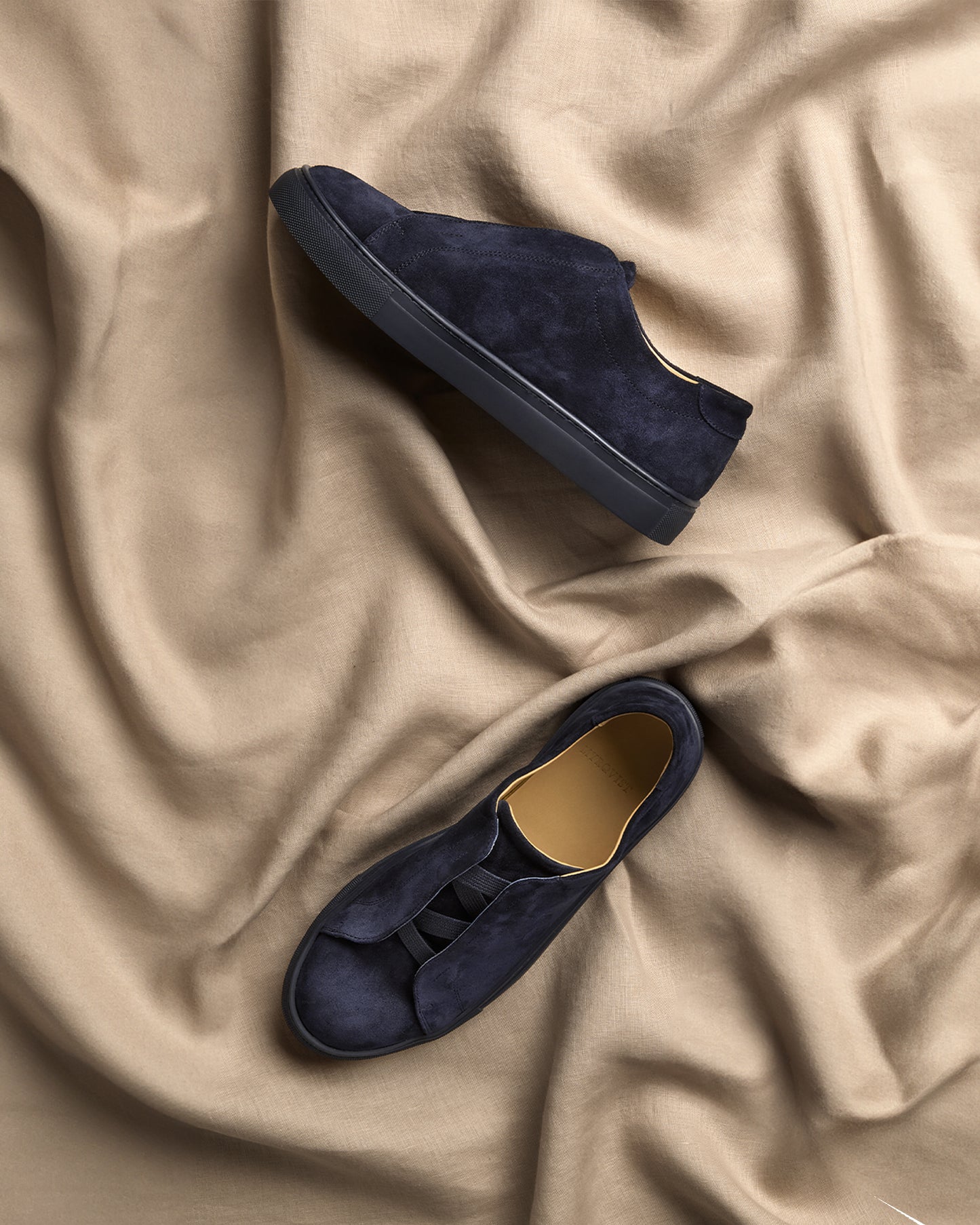 A pair of Alnö – Midnight Blue Suede sneakers artfully arranged on a draped, light brown fabric.