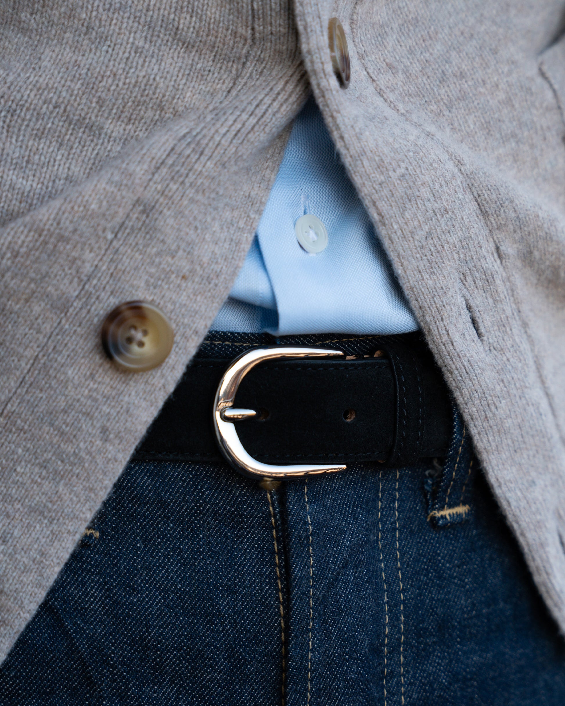 Close-up of an outfit featuring a black suede belt with a silver buckle, paired with dark denim and a beige knit cardigan.