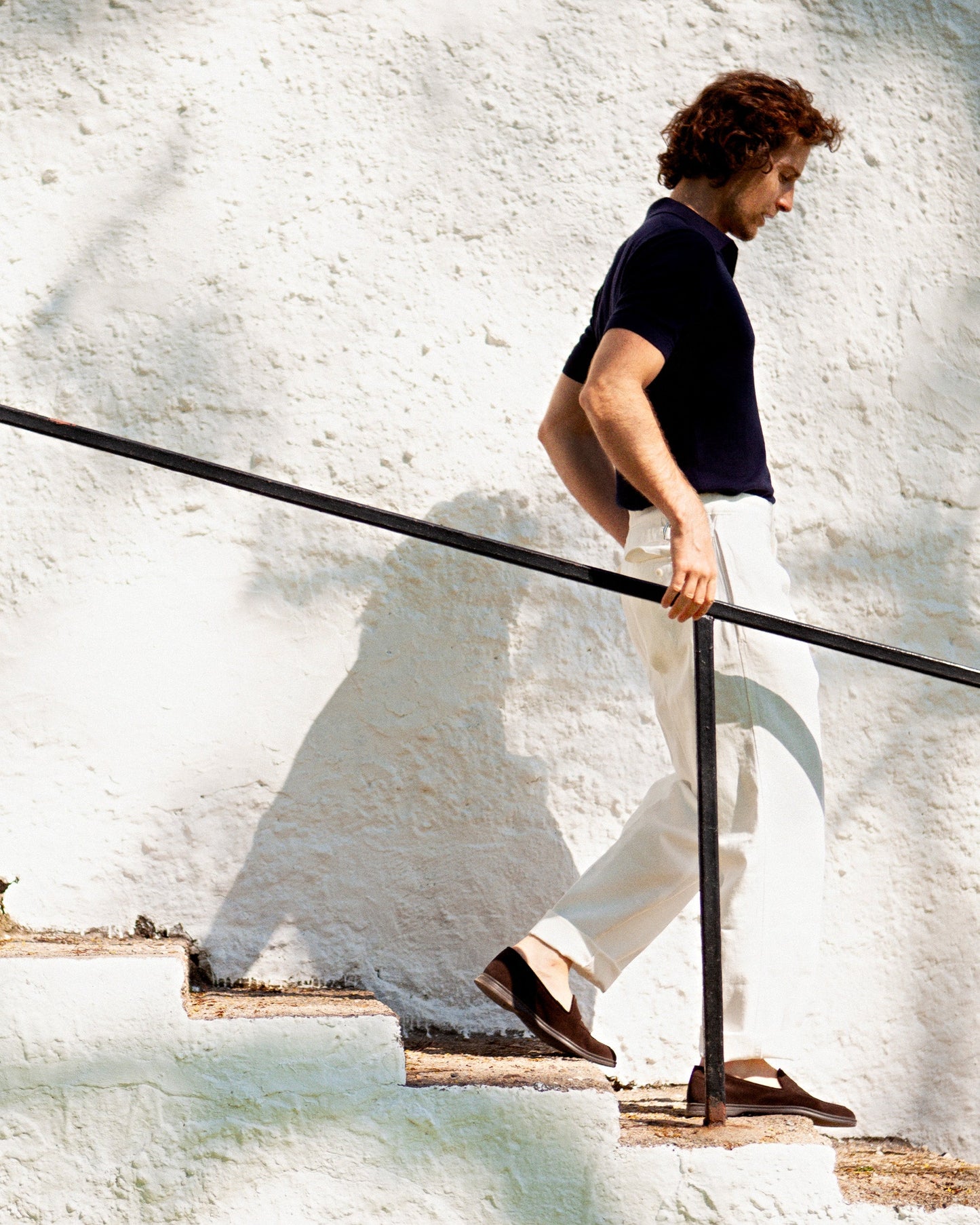 A man in a dark polo and white trousers walks down a stone staircase, wearing Brännö II – Dark Brown Suede loafers.