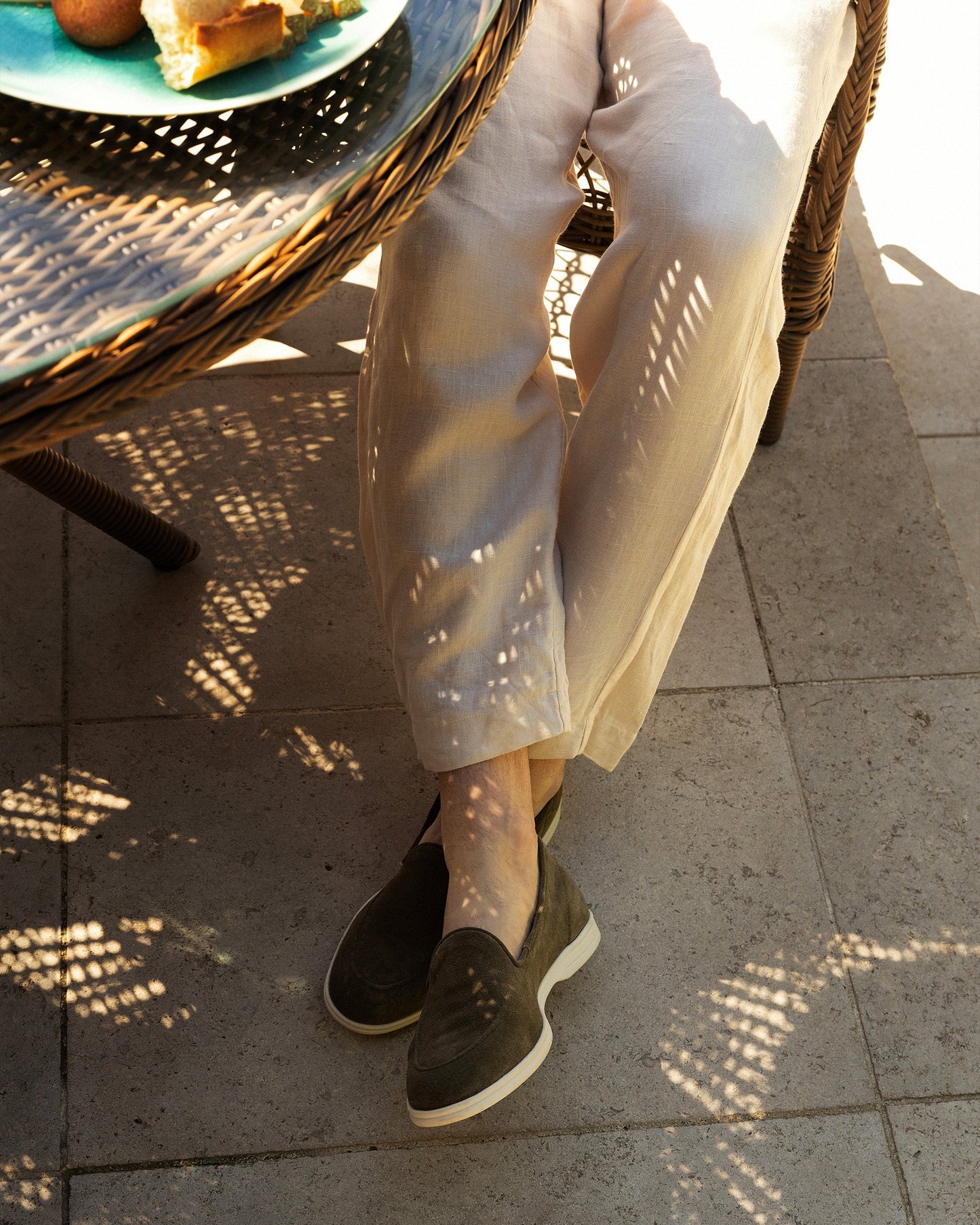 A man wearing Brännö II moss green suede loafers and linen trousers, sitting on a sunny patio.