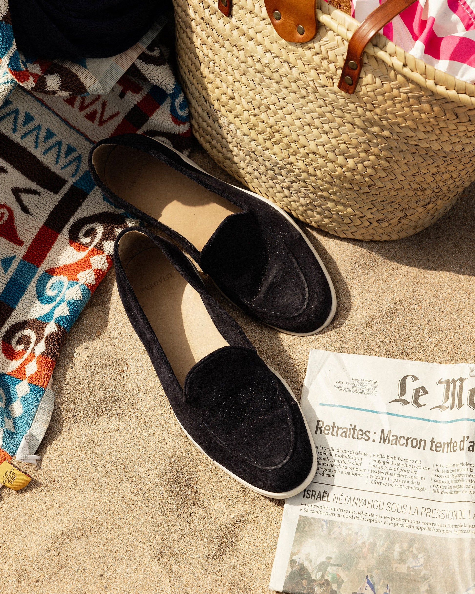 A pair of Brännö II navy suede loafers on the sand with a straw beach bag, a newspaper, and a patterned towel.