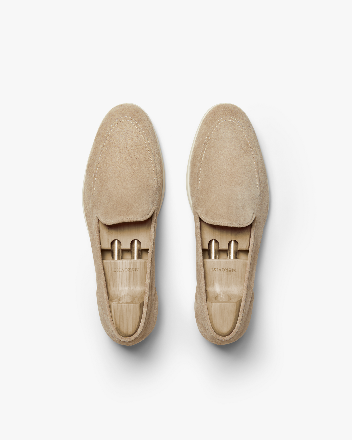 An overhead view of a pair of Brännö II – Sand Suede loafers with wooden shoe trees on a white background.