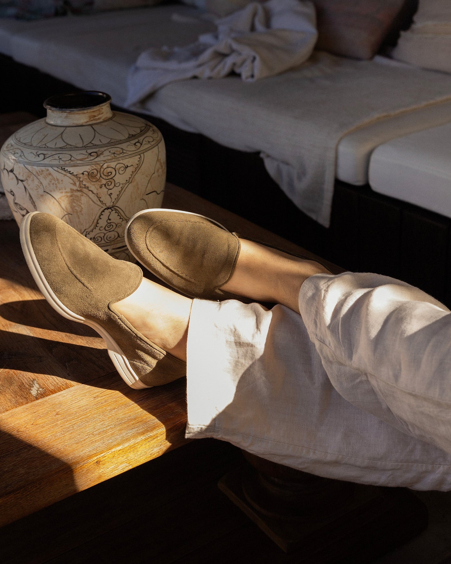 A person relaxing with their feet on a wooden table, wearing moss green suede loafers and white linen trousers.