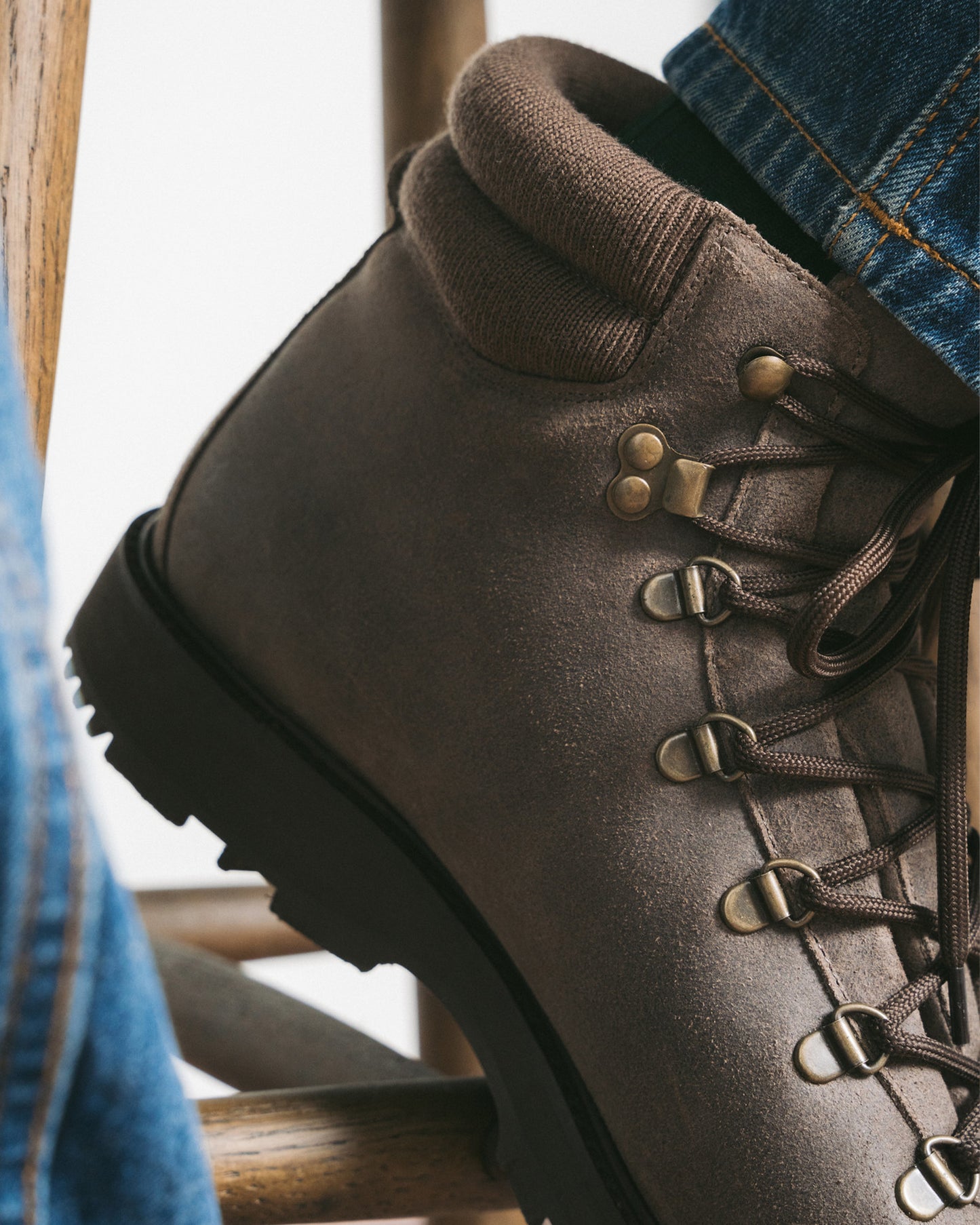 A close-up of a person wearing Duved II – Taupe Waxed Suede boots with blue jeans, resting their foot on a wooden chair.