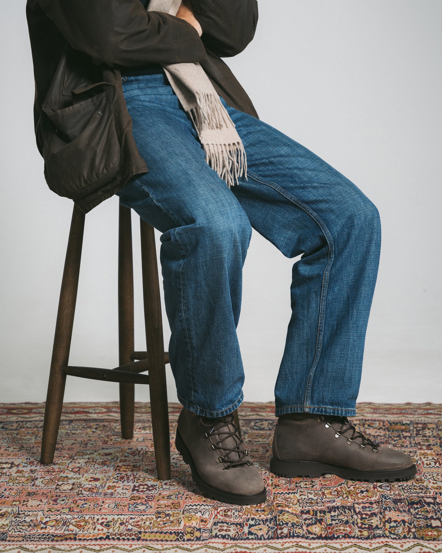 A man wearing blue jeans and a brown jacket sits on a stool, showing his Duved II – Taupe Waxed Suede boots.