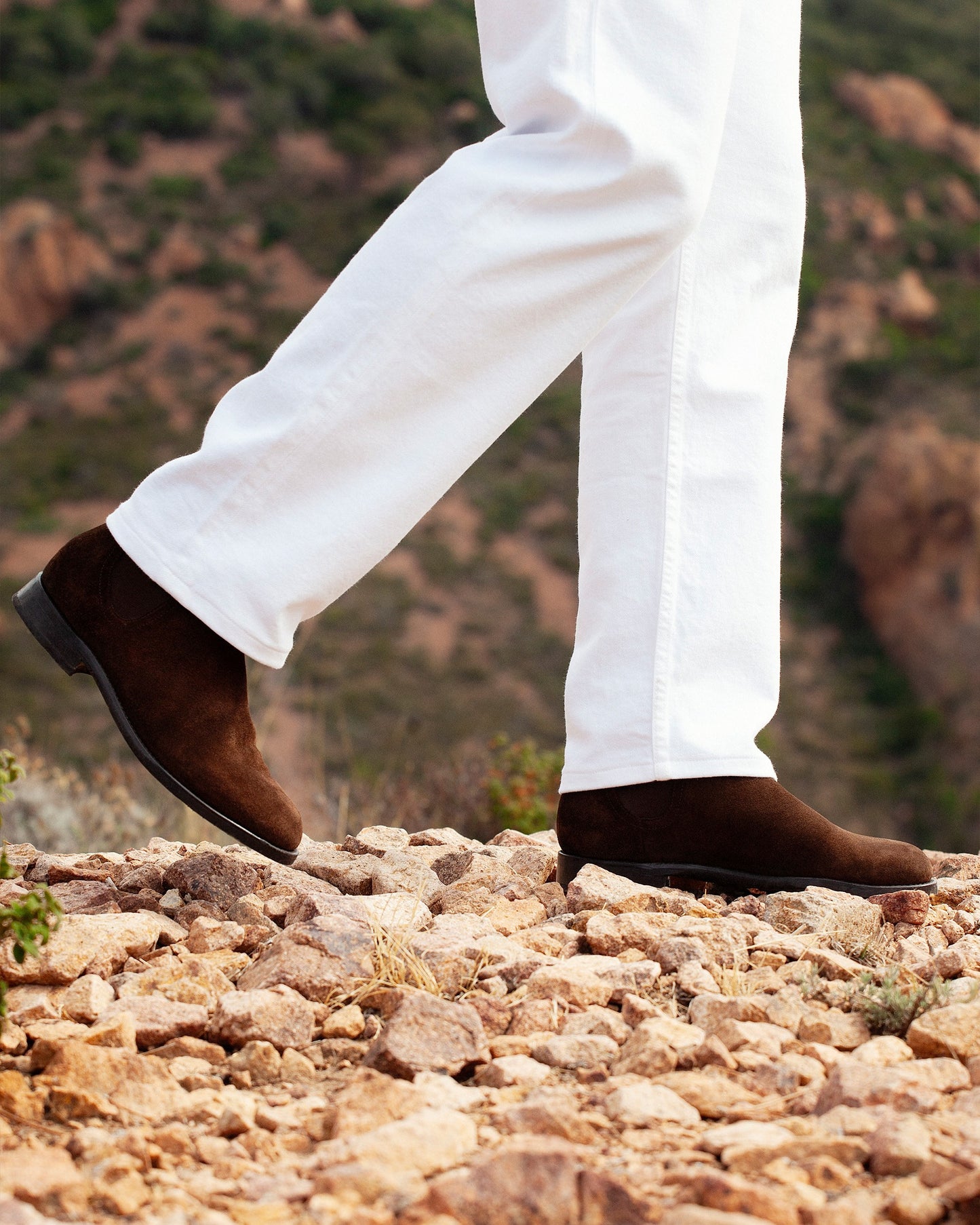 Close-up on a person wearing Elfvik – Dark Brown Suede boots with white trousers on a rocky path.