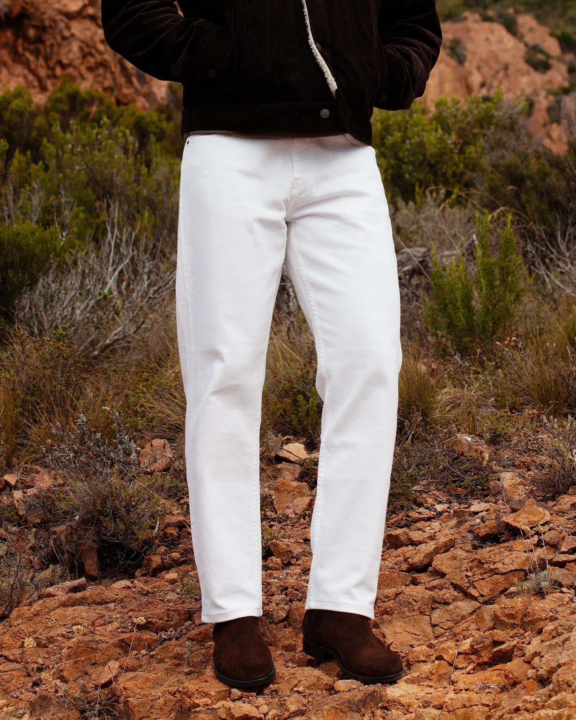 A person standing on rocky ground, wearing Elfvik – Dark Brown Suede boots with white trousers and a brown corduroy jacket.
