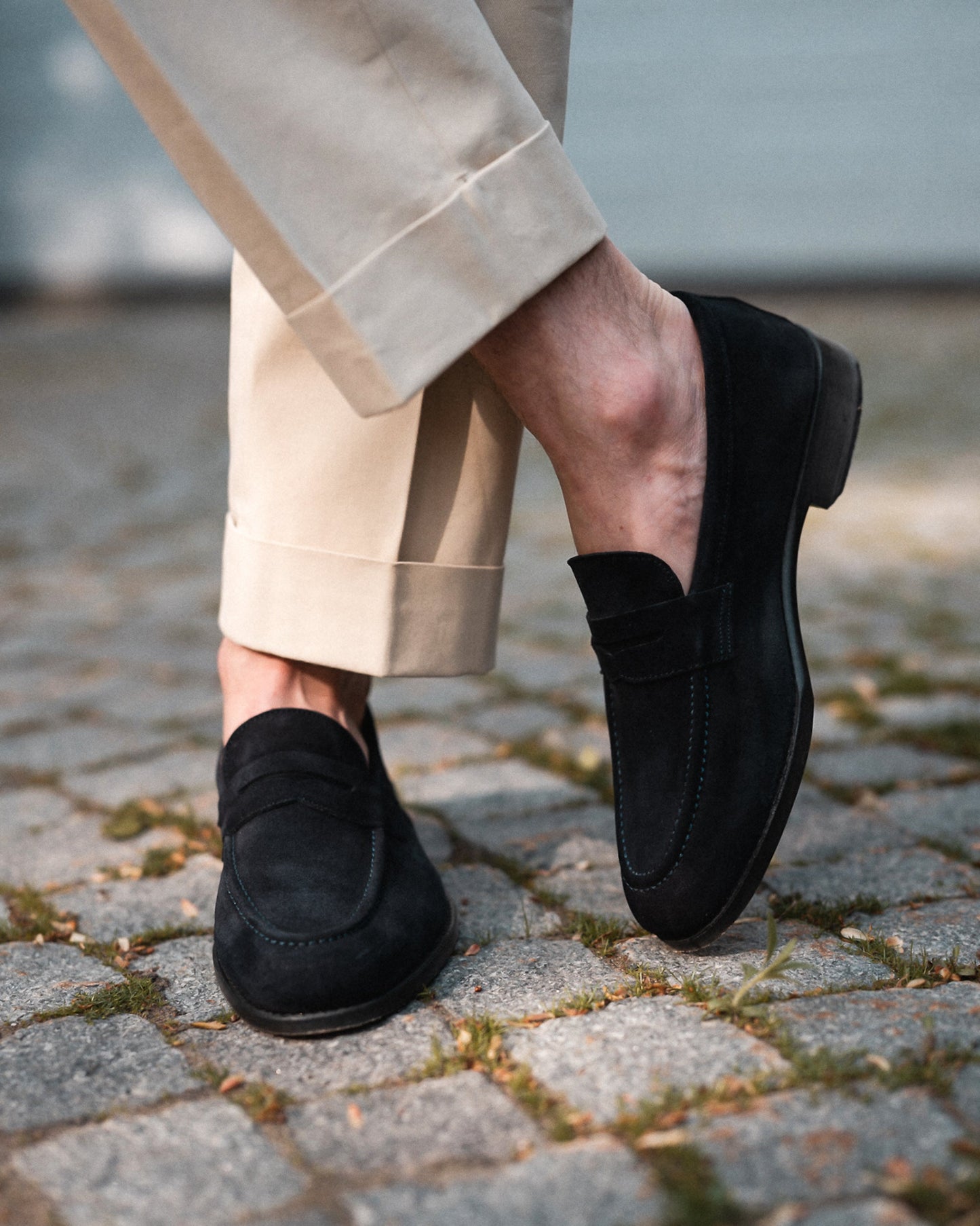 A close-up of a person wearing Lysekil II – Navy Suede loafers with light beige cuffed trousers on a cobblestone path.