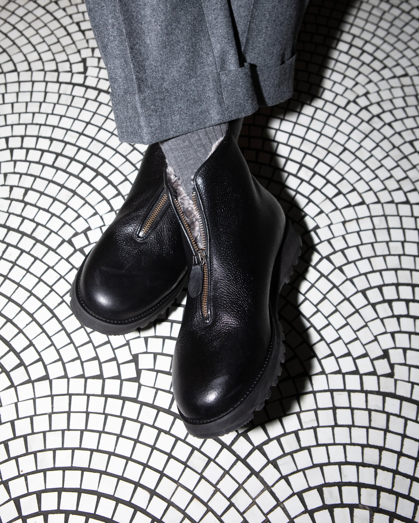 A person wearing Funäs – Black Country Calf boots with grey cuffed trousers, standing on a black and white mosaic floor.