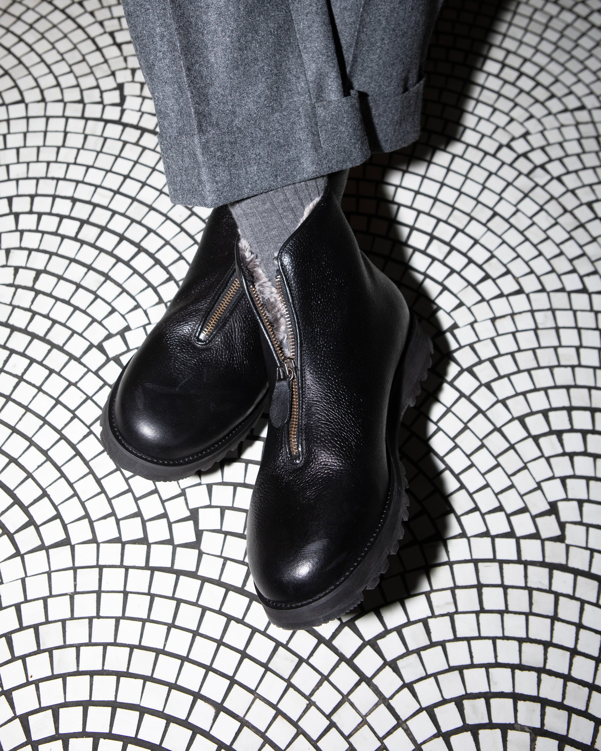 A person wearing Funäs – Black Country Calf boots with grey cuffed trousers, standing on a black and white mosaic floor.