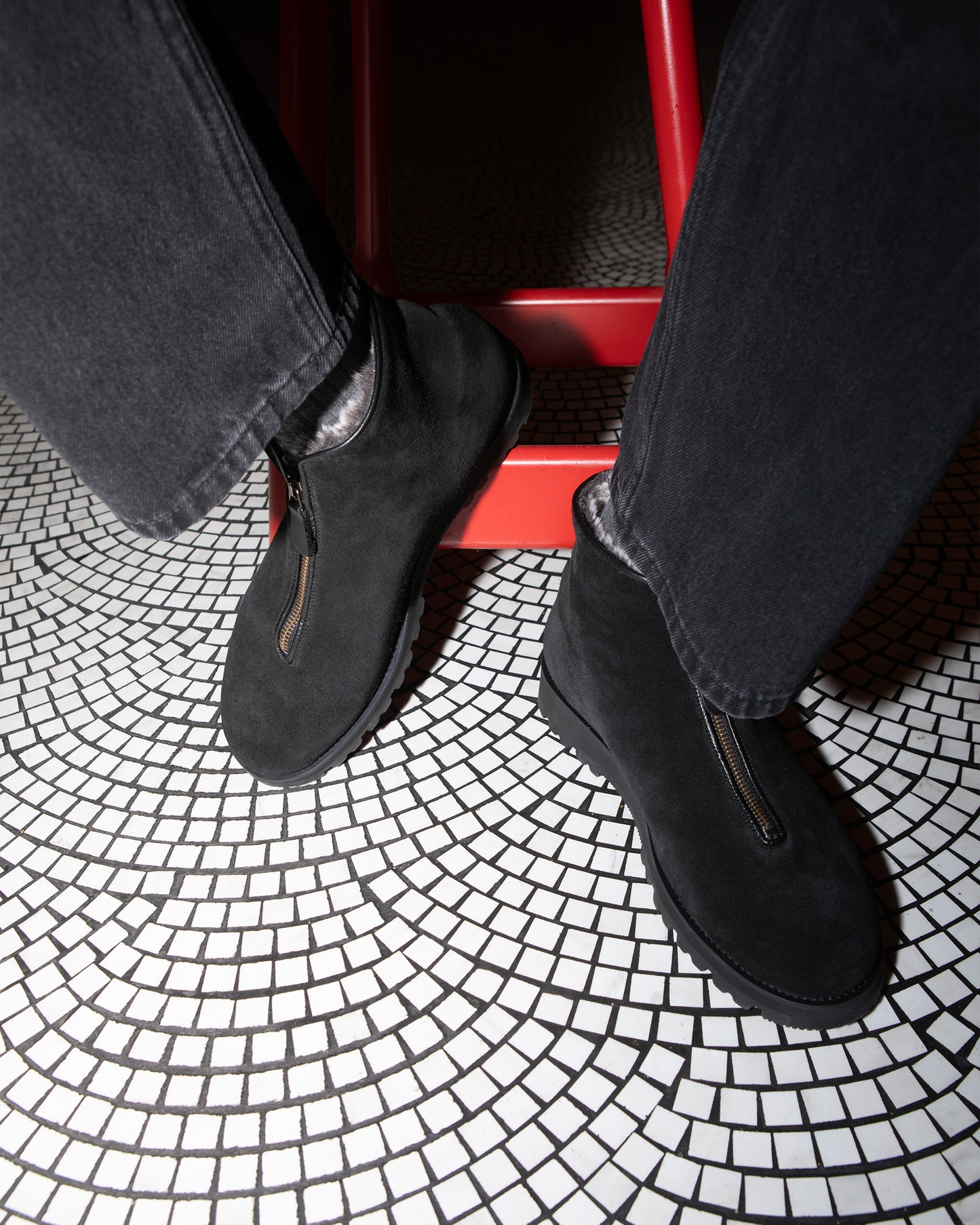A person wearing Funäs – Black Suede boots with black jeans, sitting on a red stool on a white mosaic tile floor.