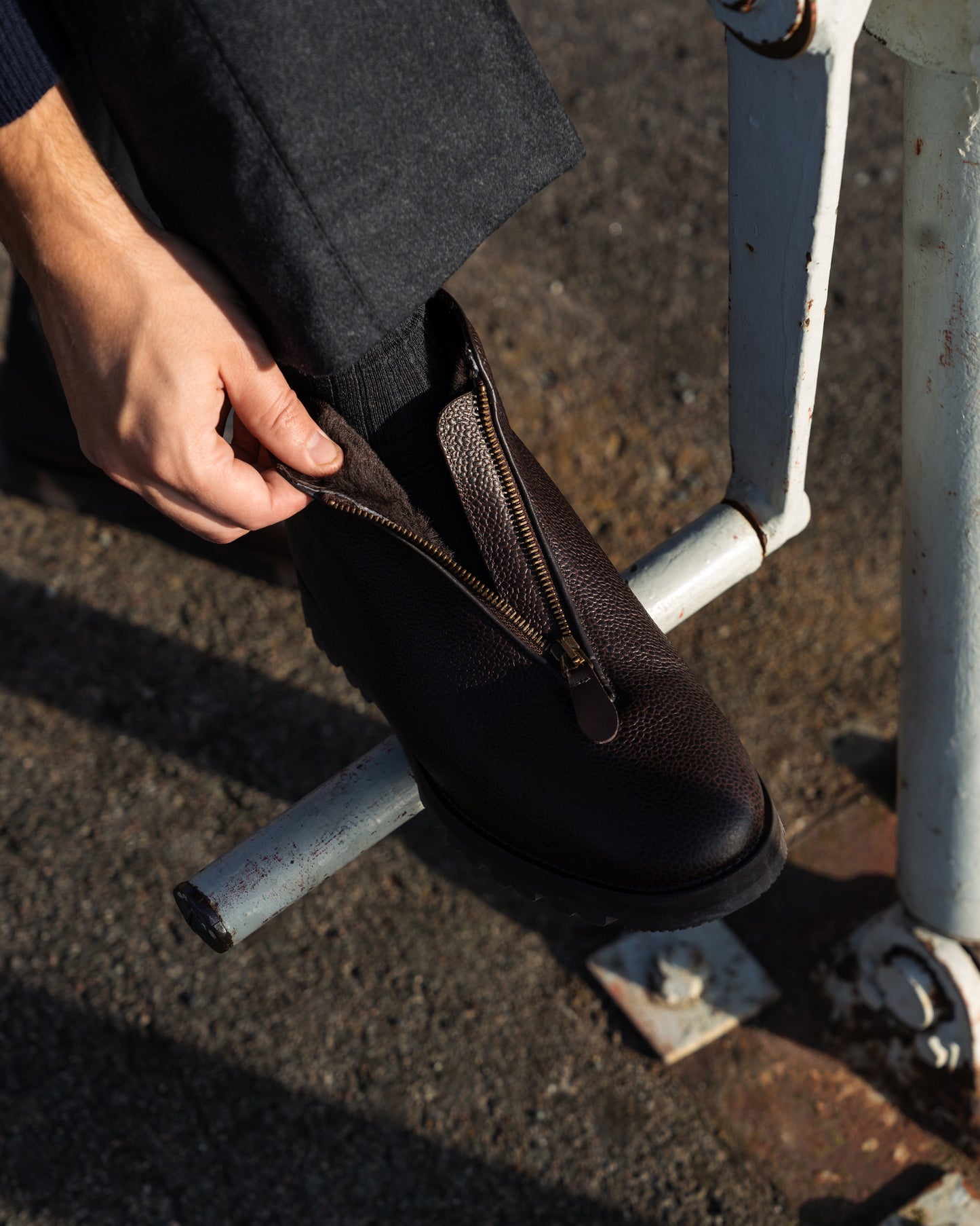 Close-up on a person opening the front zipper of a Funäs – Dark Brown Country Calf boot, worn with dark grey trousers.