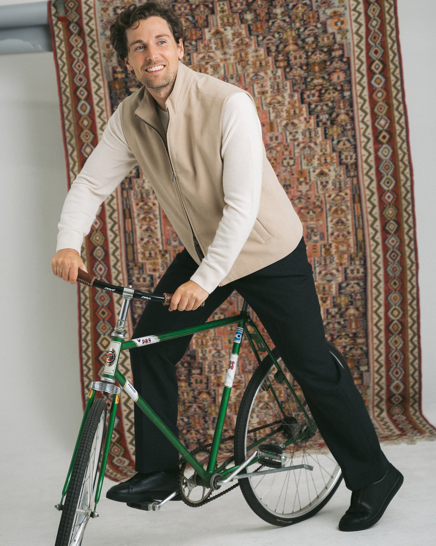 A man poses with a bicycle, wearing Alnö Shearling – Black Natural Grain sneakers with black trousers and a beige vest.