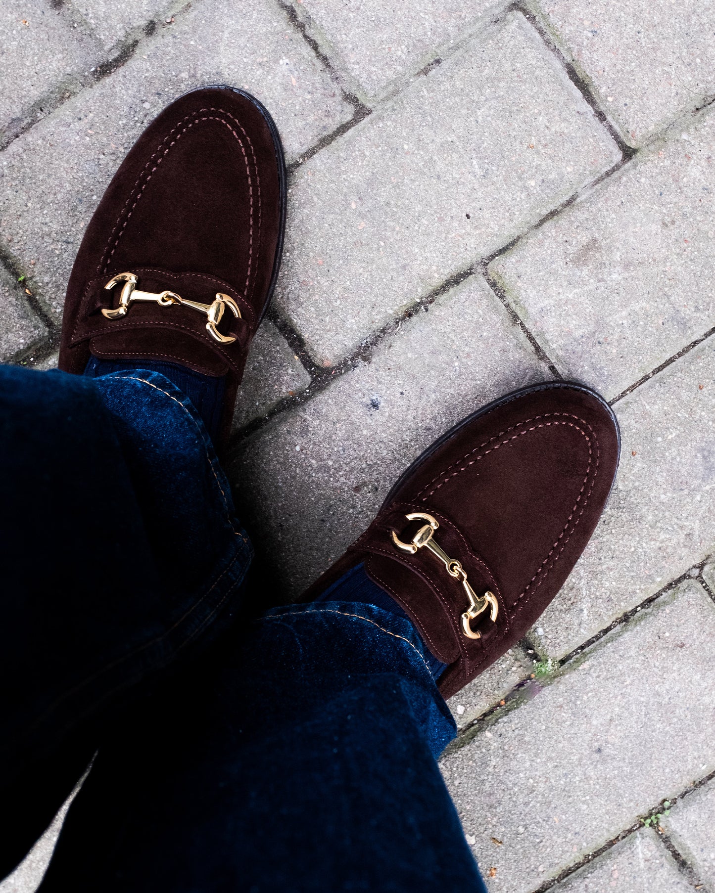 Karlberg loafers in dark brown suede worn with dark denim, featuring gold horsebit detail and fine stitching, shown on a stone pavement for a relaxed yet refined look.