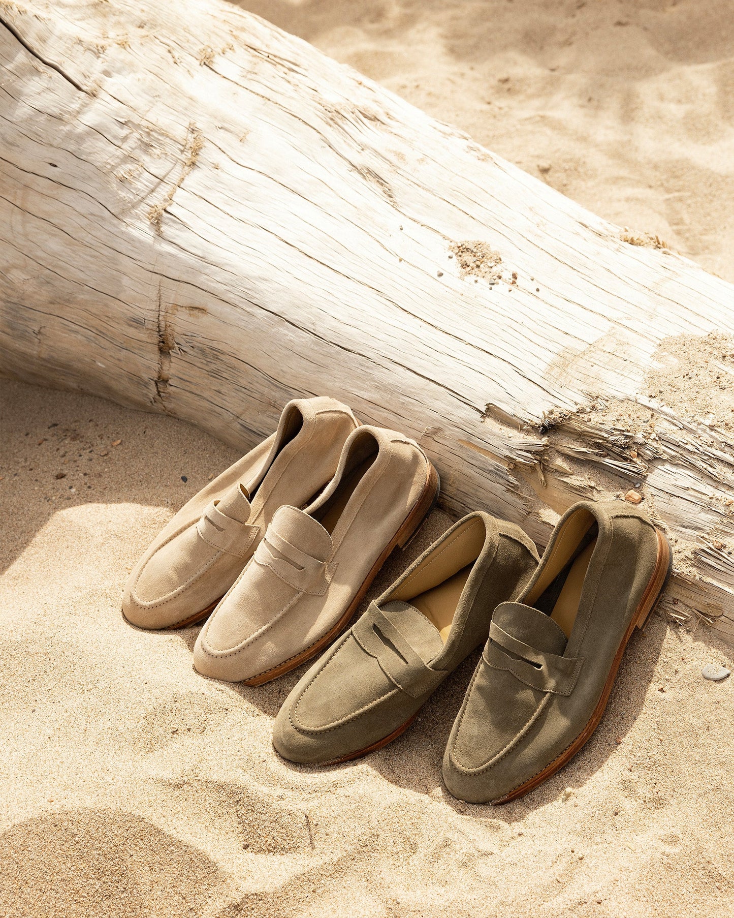 Lysekil II loafers in green and sand suede resting on a beach against a piece of driftwood.