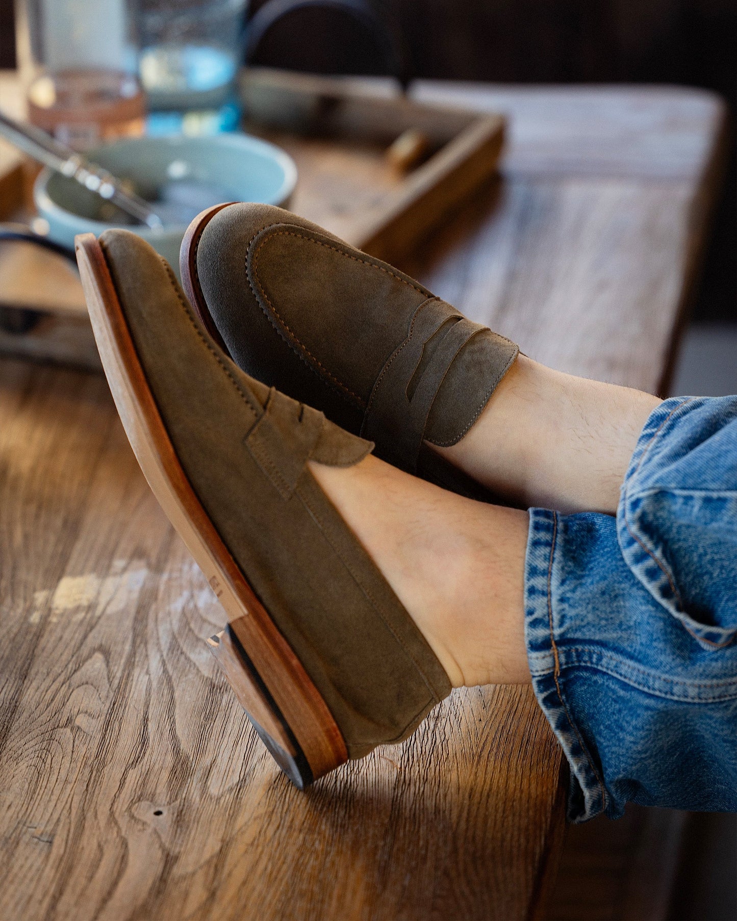A lifestyle shot of a person wearing green suede penny loafers and jeans, relaxing with their feet on a wooden table.
