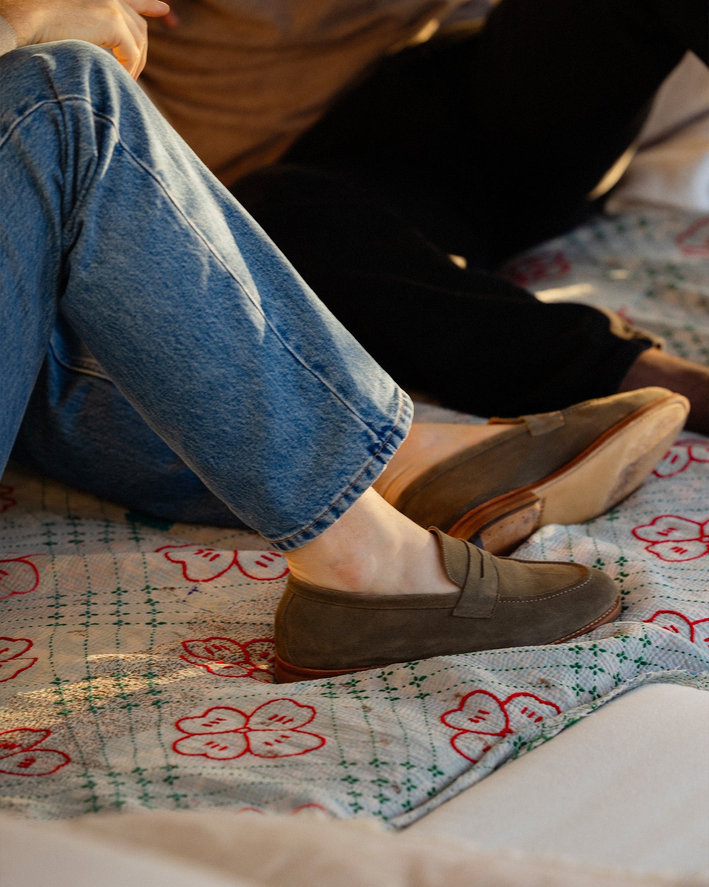 A person wearing Lysekil II green suede loafers and blue jeans, relaxing on a patterned quilt.