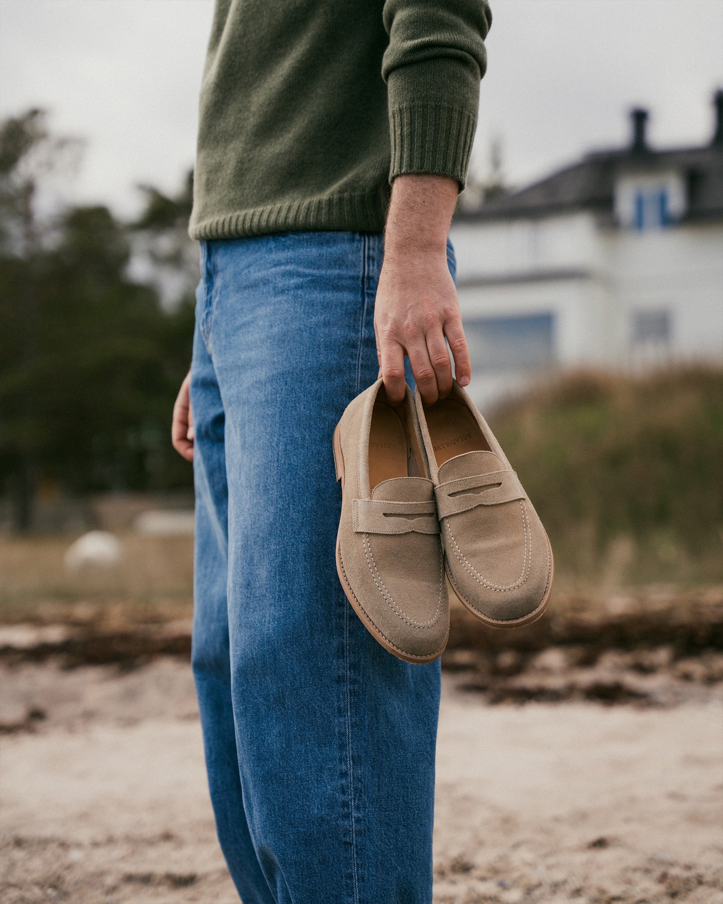 Close-up of a man in a green sweater and blue jeans holding Lysekil II – Sand Suede loafers on a beach.