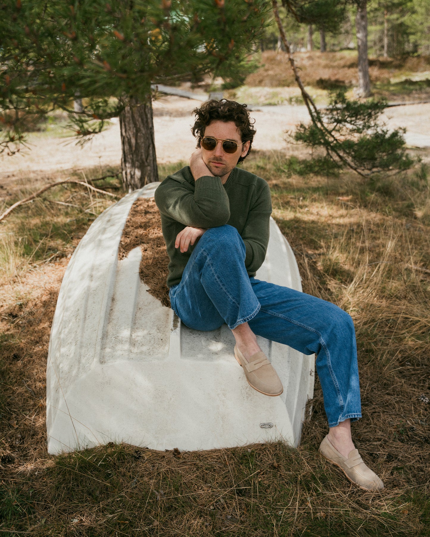 Man in a green sweater and blue jeans relaxes on an overturned boat, wearing Lysekil II – Sand Suede loafers.