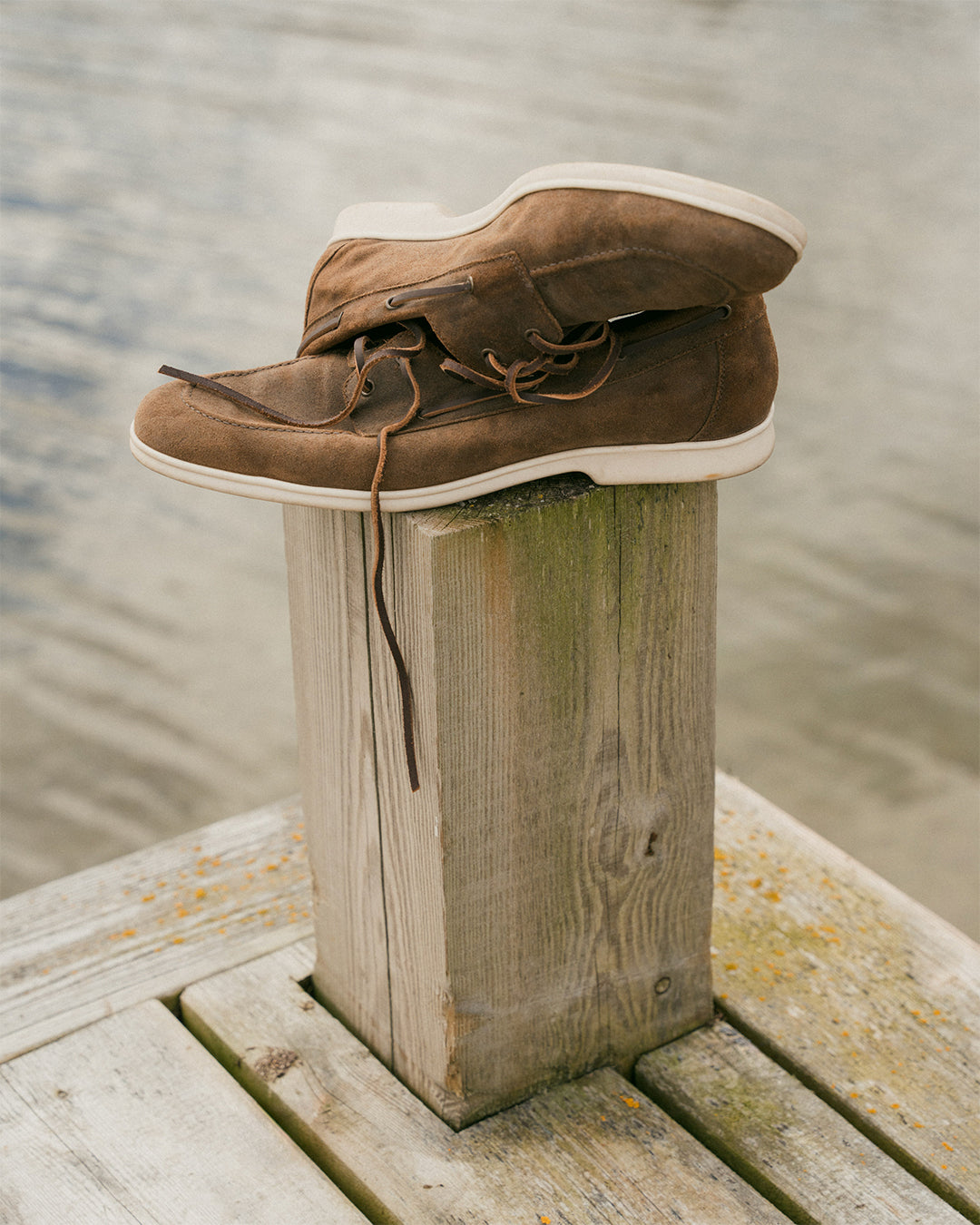 Sandhamn – Chestnut Waxed Suede boat shoe rests on a weathered wooden post on a pier by the water.