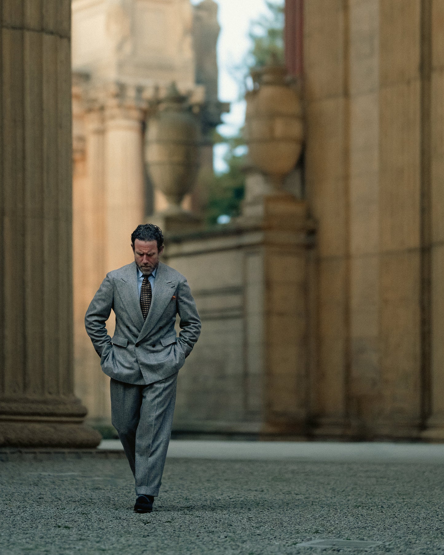 A man walks in a courtyard with large columns, wearing a grey plaid suit paired with Mölle II – Black Suede loafers.