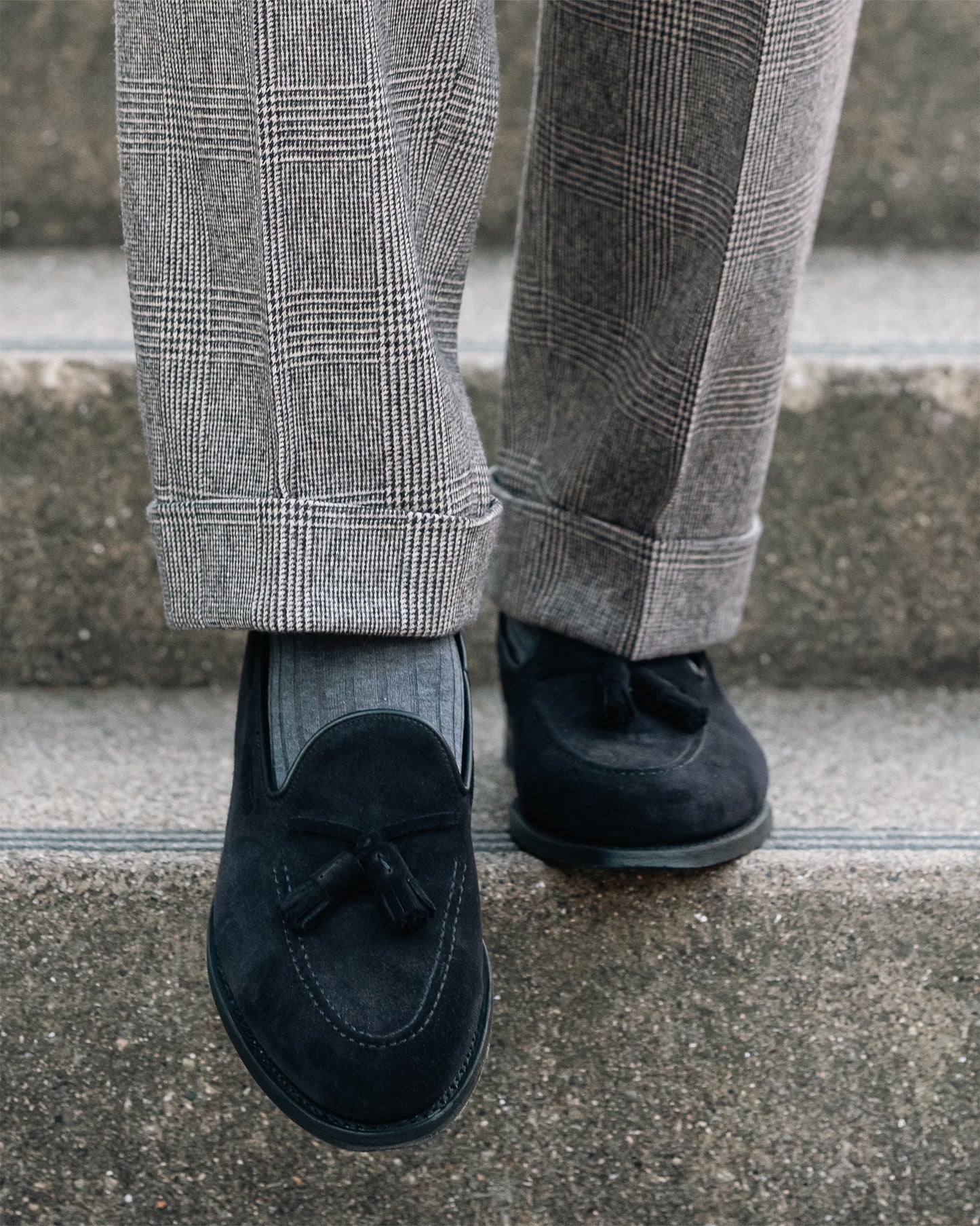 A close-up of Mölle II – Black Suede loafers paired with grey socks and cuffed plaid trousers on concrete steps.