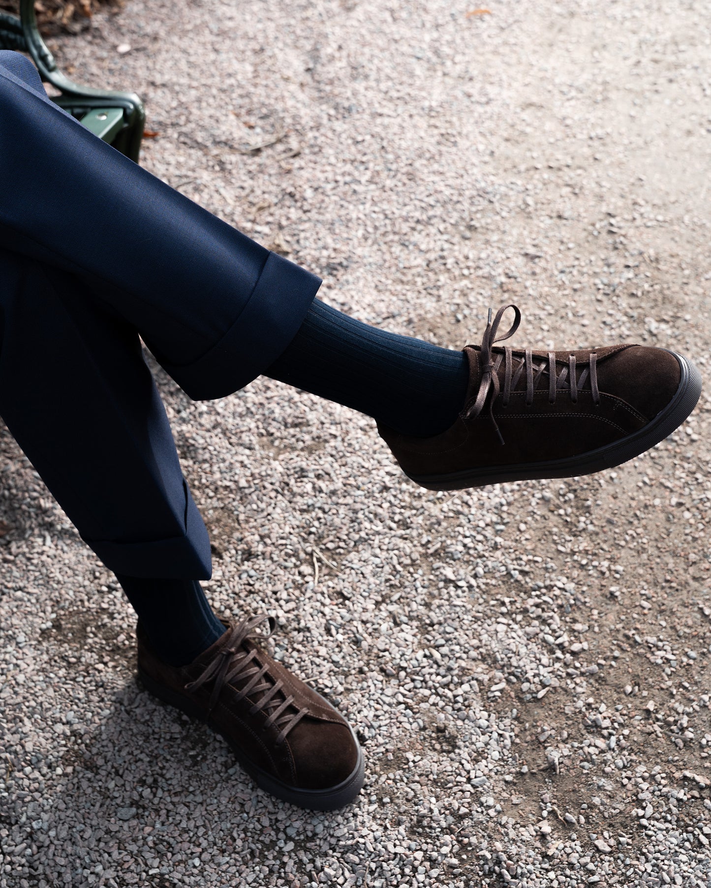 A person sitting on a park bench wearing Oaxen Monochrome II – Dark Brown Suede sneakers with cuffed navy trousers.