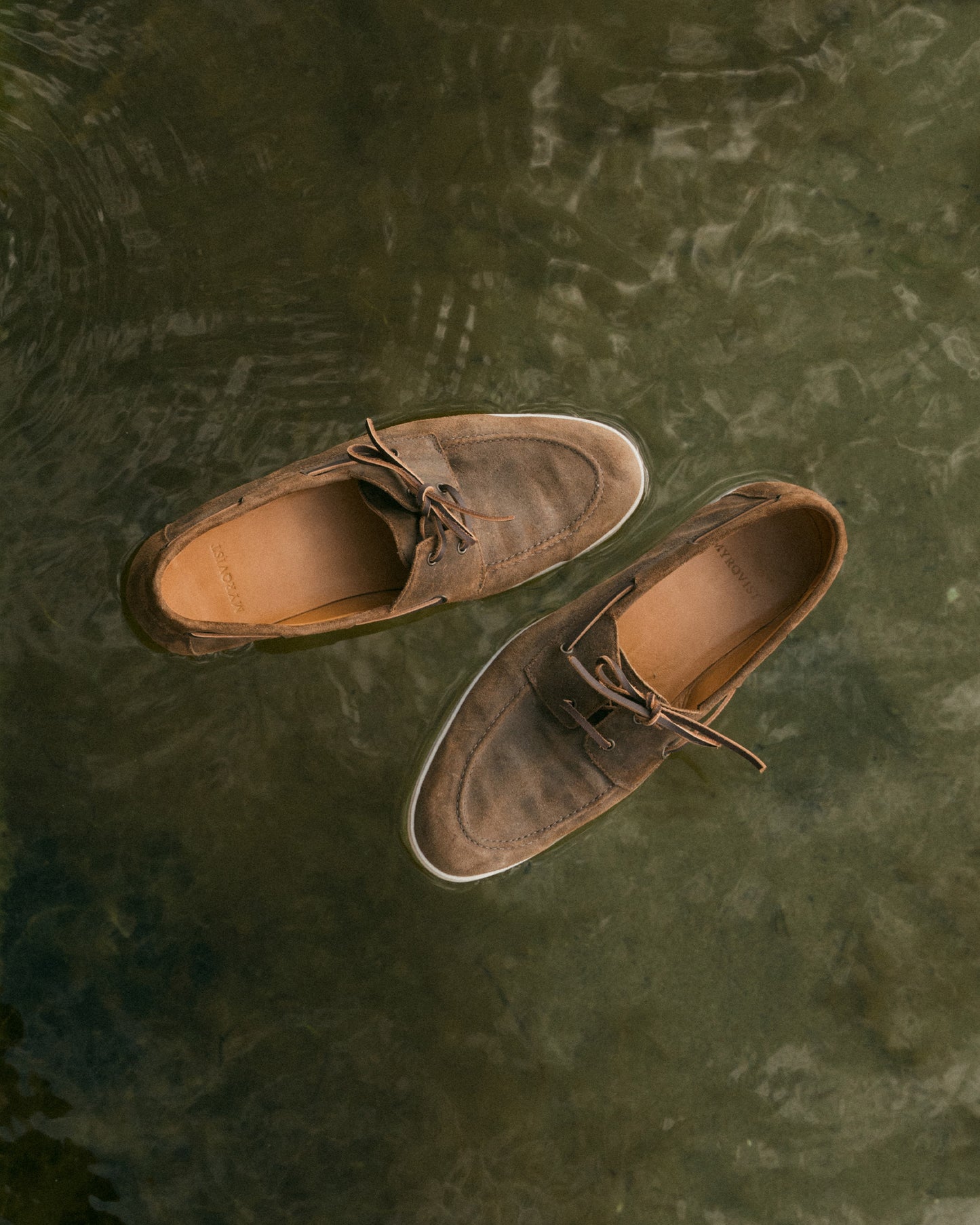 A pair of Sandhamn – Chestnut Waxed Suede boat shoes seen from above, floating on the surface of dark, rippling water.