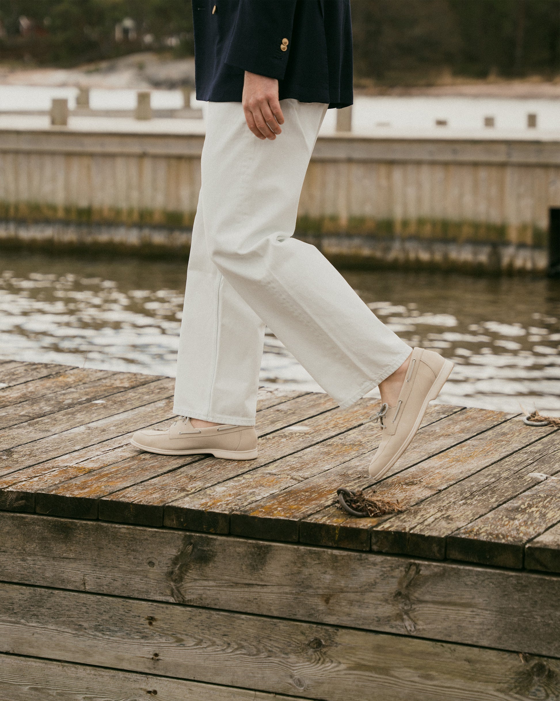 Close-up of a person walking on a wooden dock, wearing Sandhamn – Beige Nubuck boat shoes with white trousers.