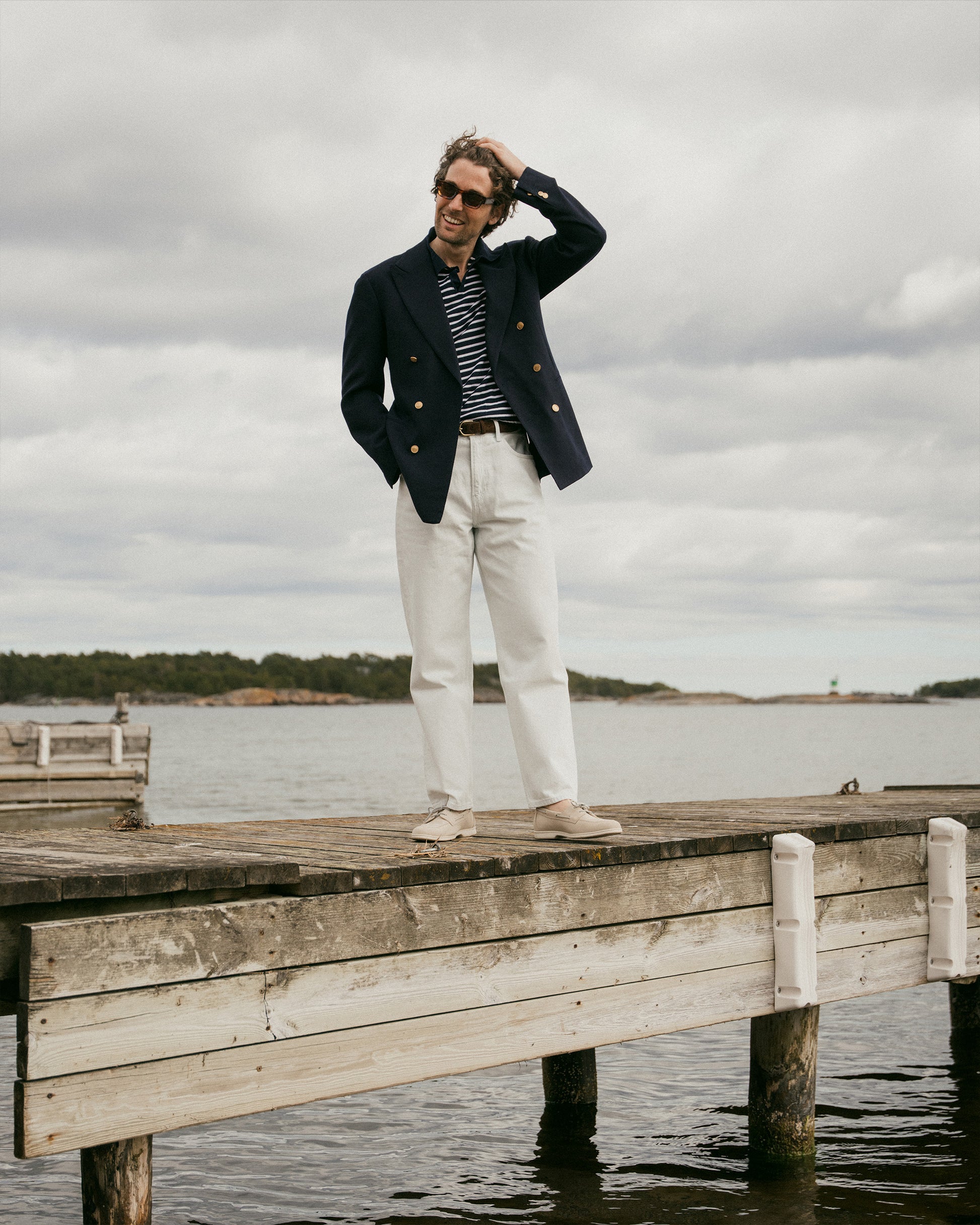 A man on a wooden pier wearing a navy blazer, light trousers, and Sandhamn – Beige Nubuck boat shoes.