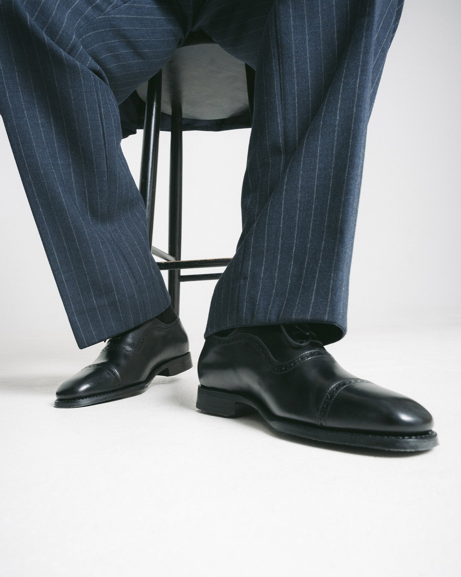 A person seated wearing navy pinstripe trousers and Skytteholm – Black Calf oxford shoes.