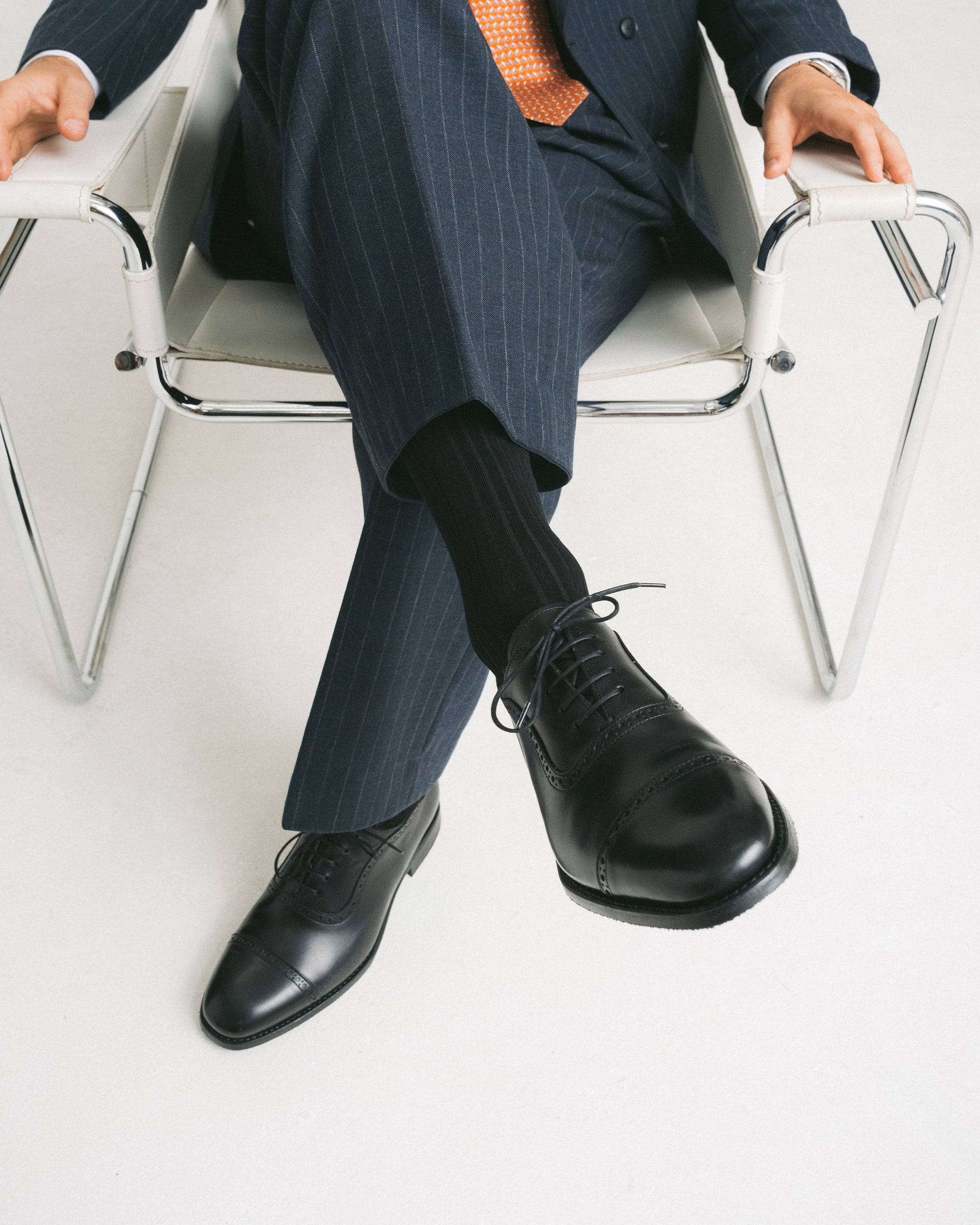 A man seated in a modern chair wearing a pinstripe suit, with a focus on his Skytteholm Black Calf oxford shoes.