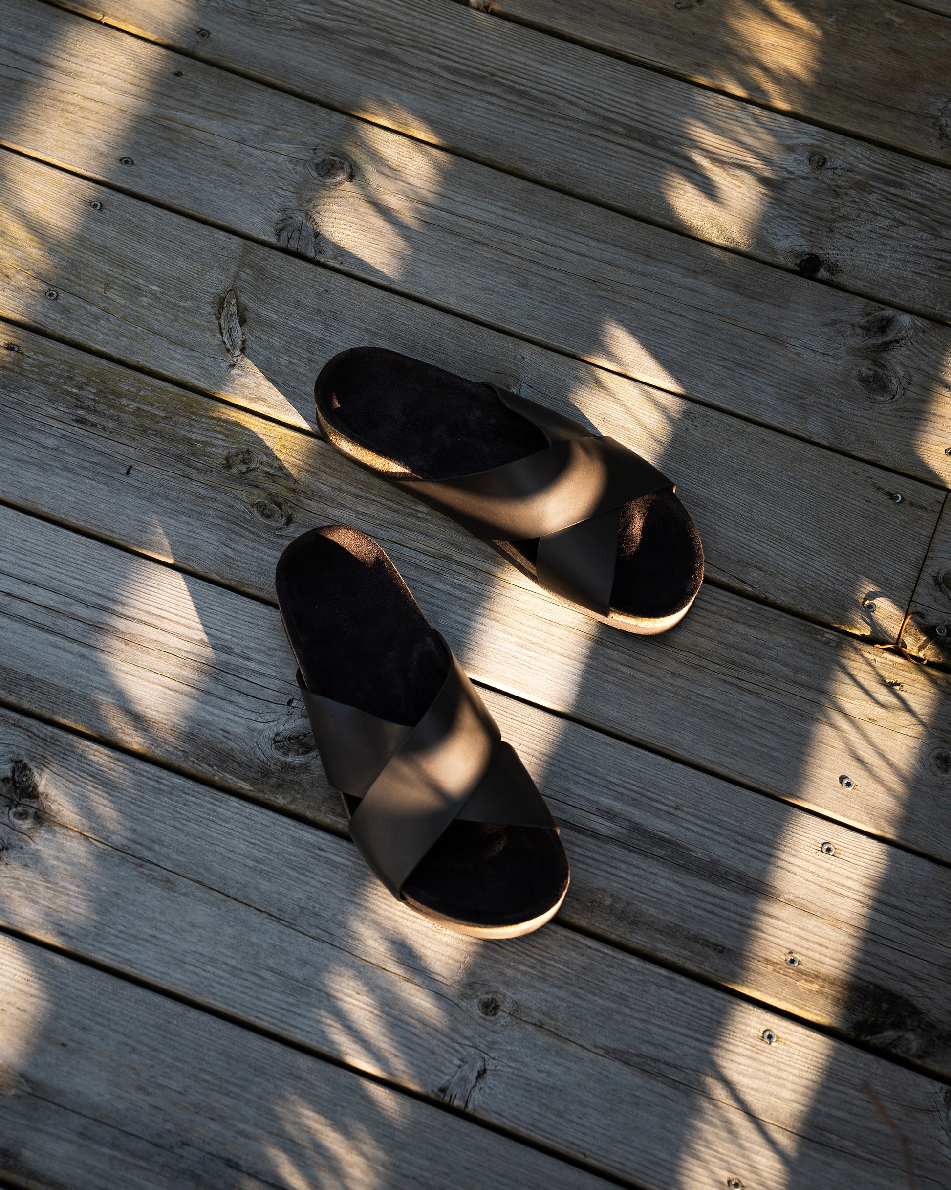 An overhead view of Solvik dark brown calf sandals on a rustic wooden deck, with strong sunlight creating long shadows.