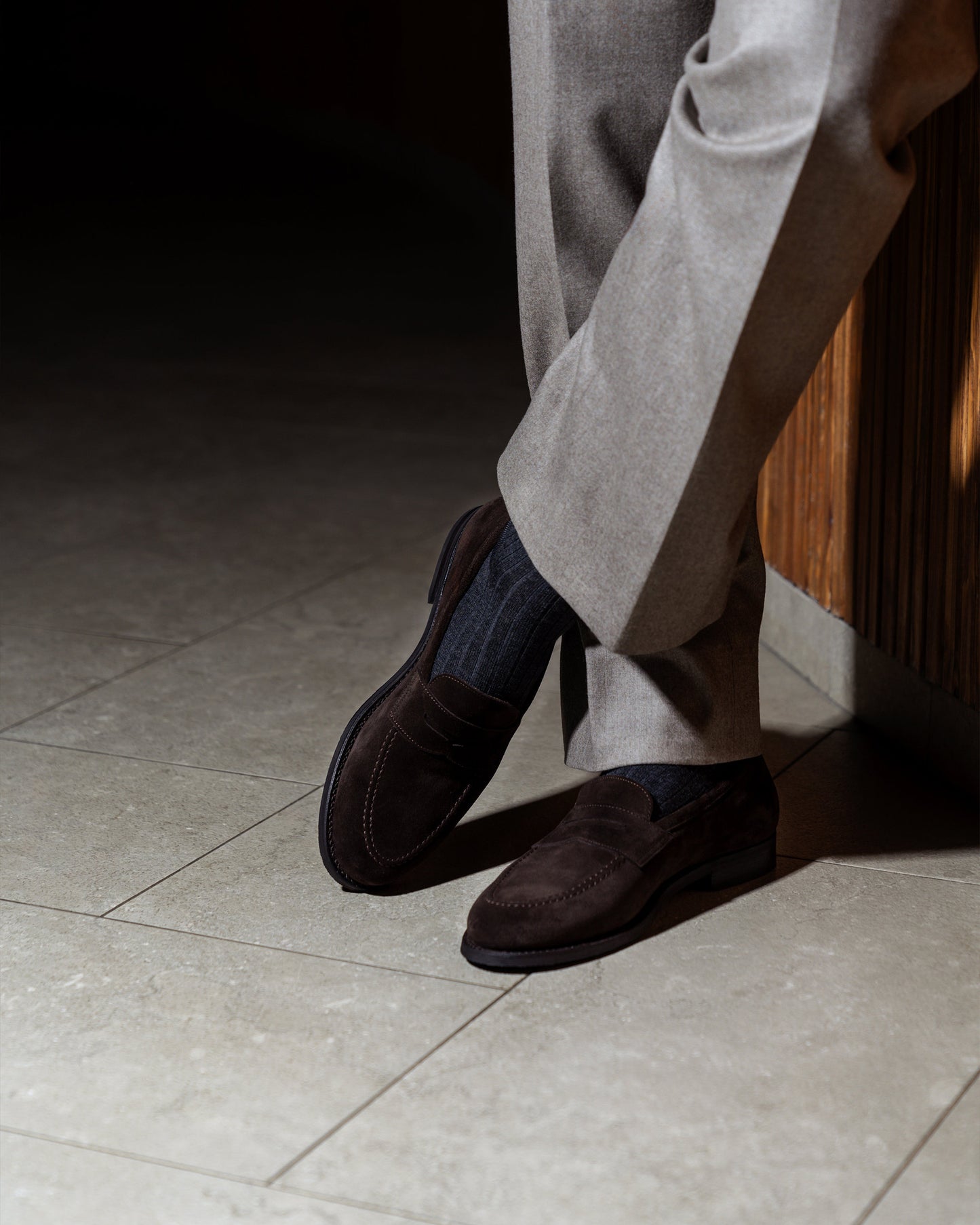 A person with legs crossed wearing Stenhammar II – Dark Brown Suede loafers with light brown trousers and dark grey socks.