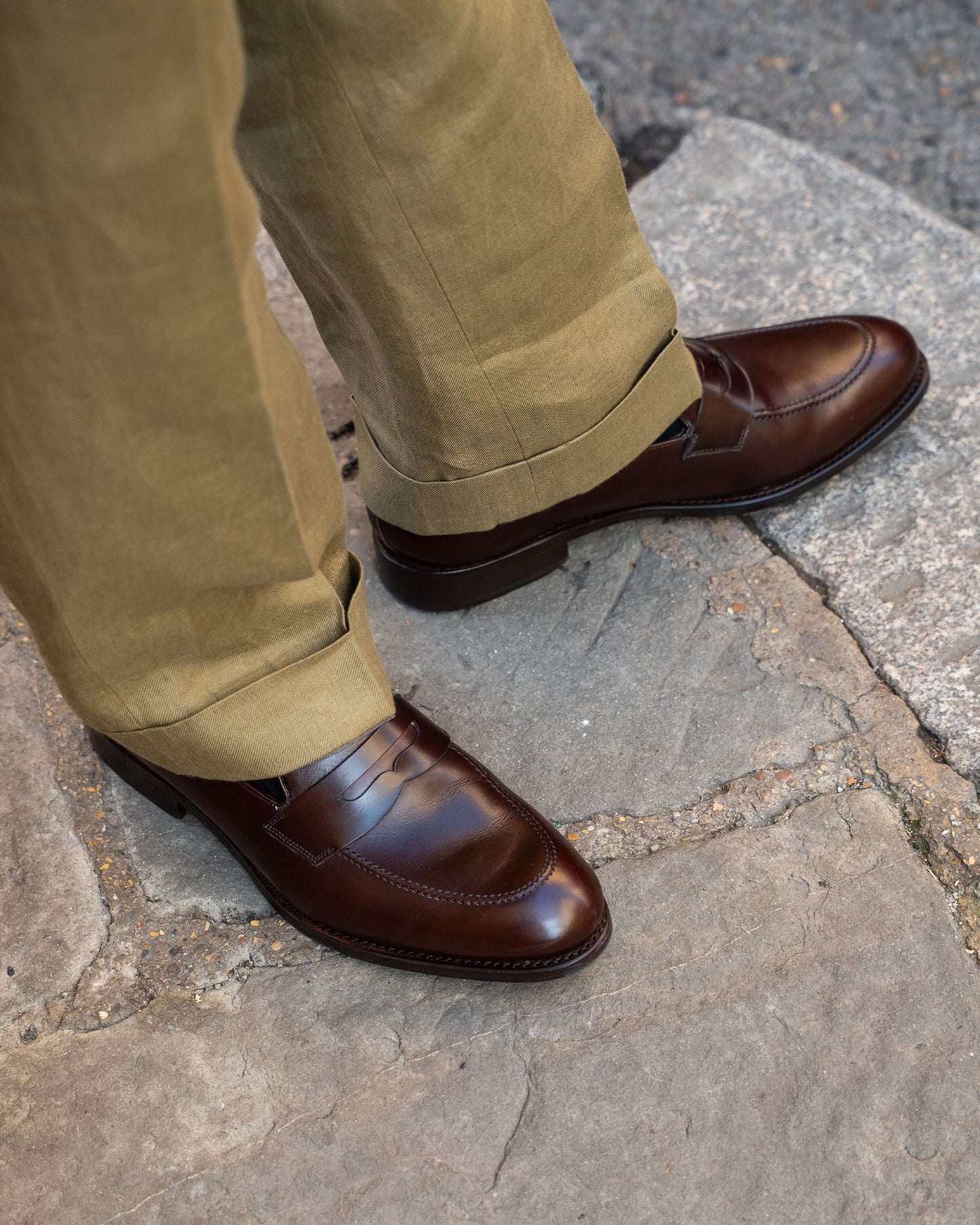 Close-up on a person wearing Stenhammar II – Dark Brown Calf loafers with cuffed olive green trousers on a stone street.