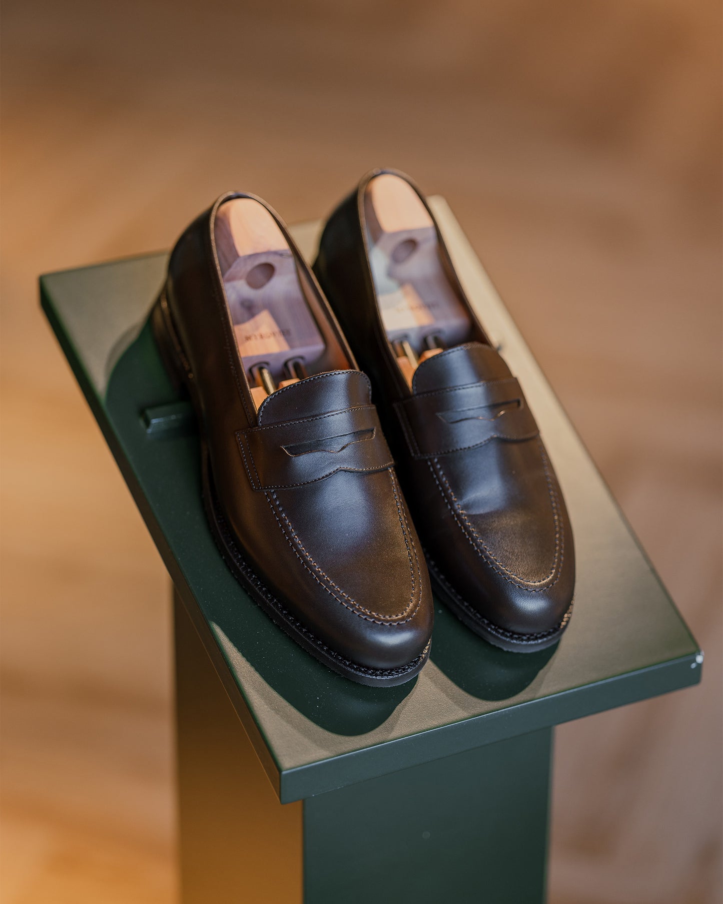 A pair of Stenhammar II – Dark Brown Calf penny loafers with wooden shoe trees displayed on a green stand.