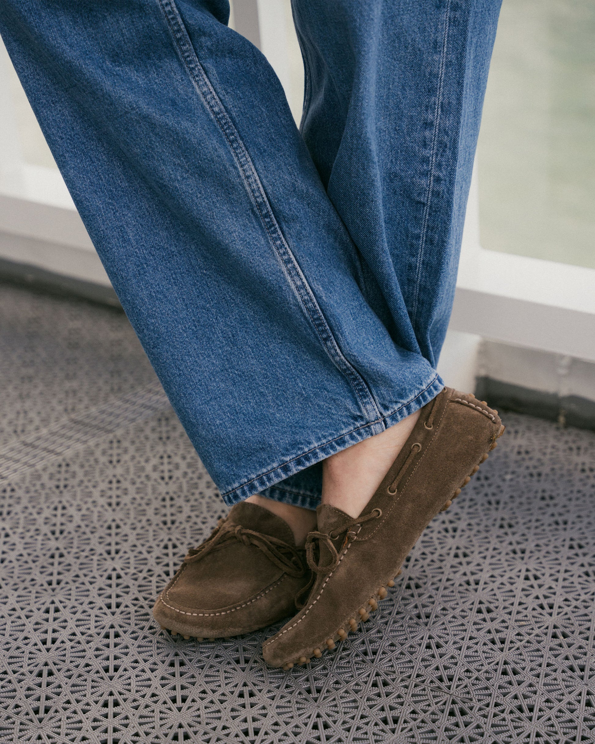 Close-up of a person wearing Tofta – Taupe Suede driving shoes with blue jeans, with their feet crossed.